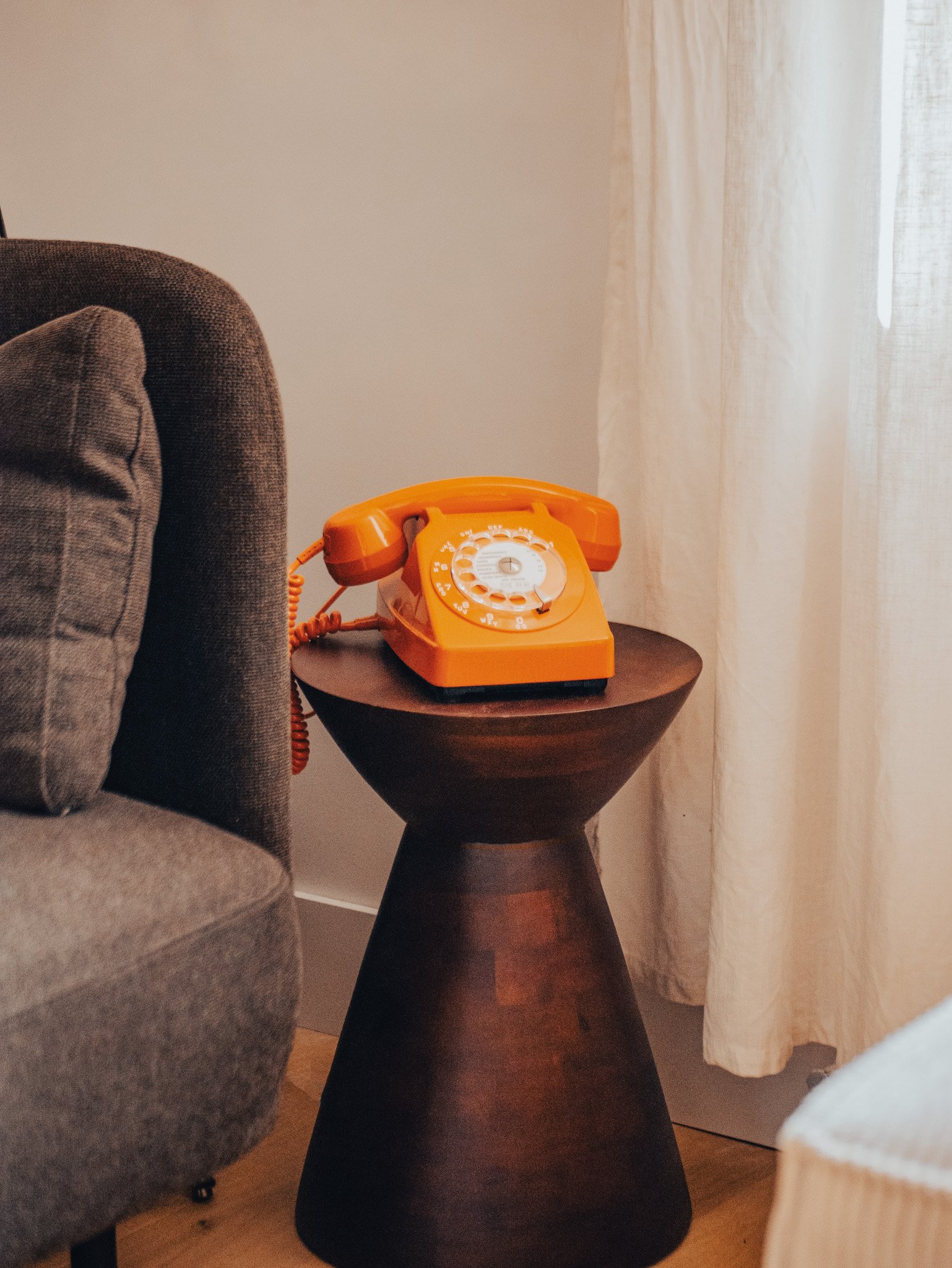 Un téléphone fixe orange posé sur une petite table en bois sombre, à côté d'un canapé gris dans une pièce lumineuse avec rideaux blancs.