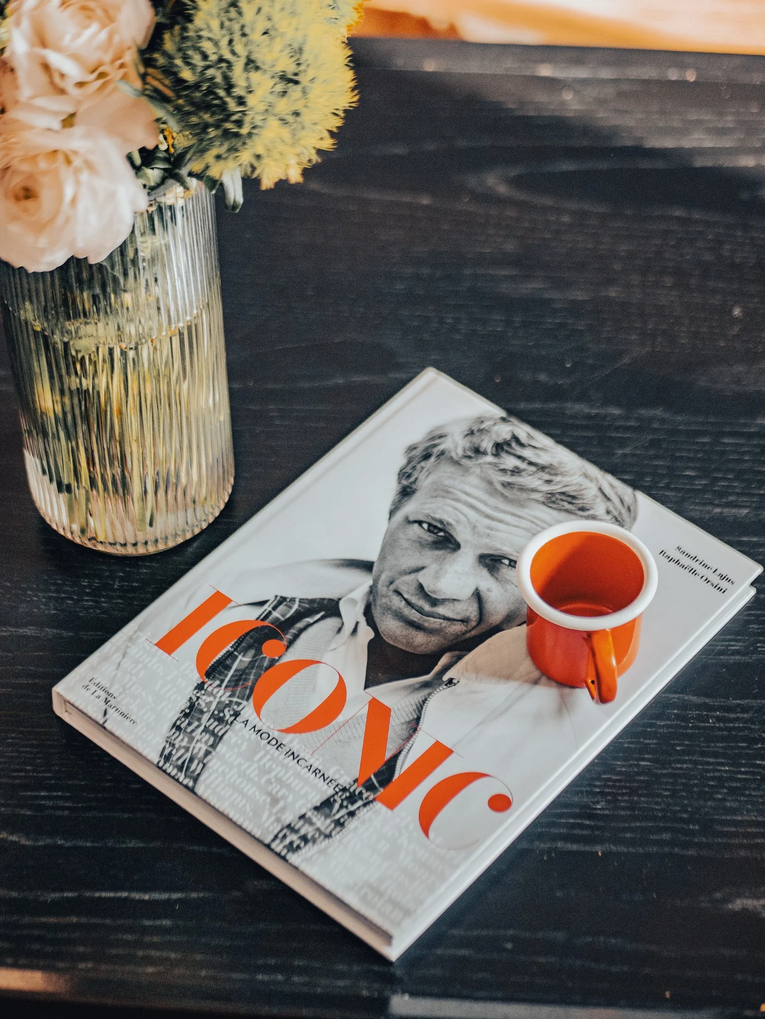 Un livre avec la couverture montrant une photo en noir et blanc d'un homme portant une chemise. Sur le livre est posé une petite tasse orange, un mug. Une plante avec des fleurs blanches et jaunes dans un vase en verre rayé de vertical.