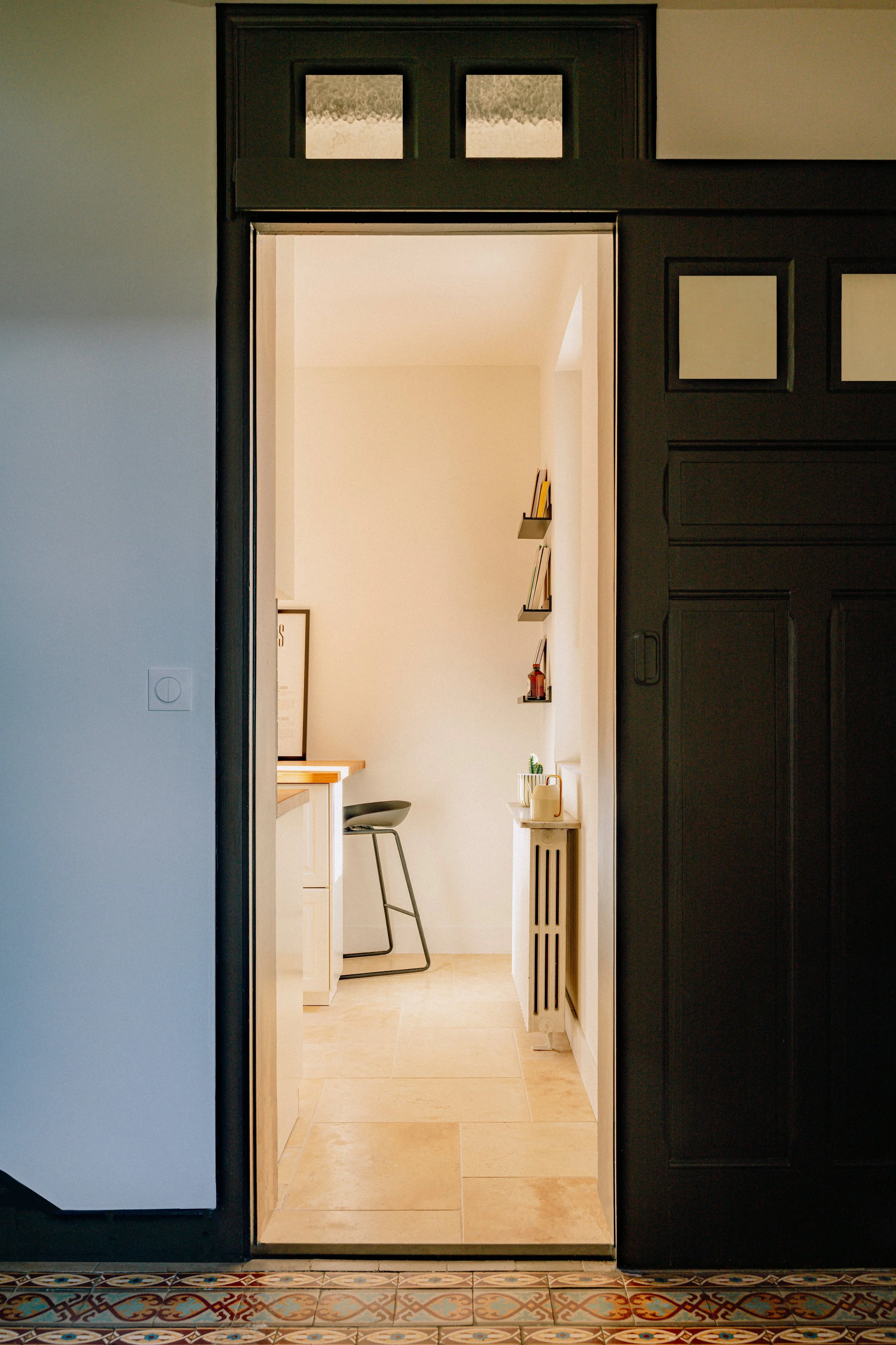 Vue d'une porte ouverte menant à une cuisine lumineuse avec des étagères contenant des livres et des décorations, une chaise de bar, un radiateur blanc et un sol en carreaux beige. Le seuil est en mosaïque colorée.