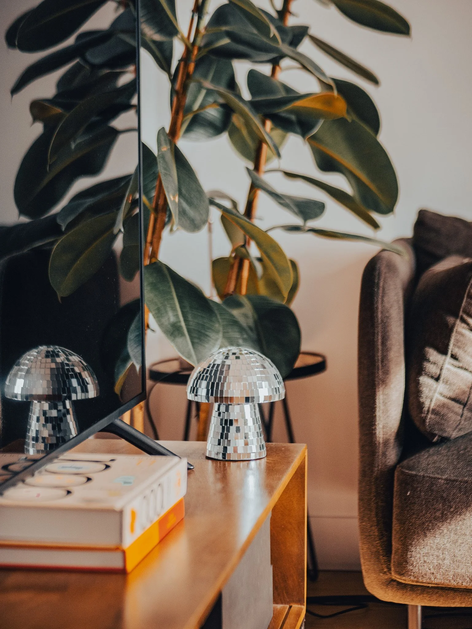 Une lampe décorative en forme de champignon recouverte de petits miroirs posée sur une table en bois, une télévision à gauche, un grand feuillage d'une plante verte derrière, un canapé beige avec un coussin patches à droite.