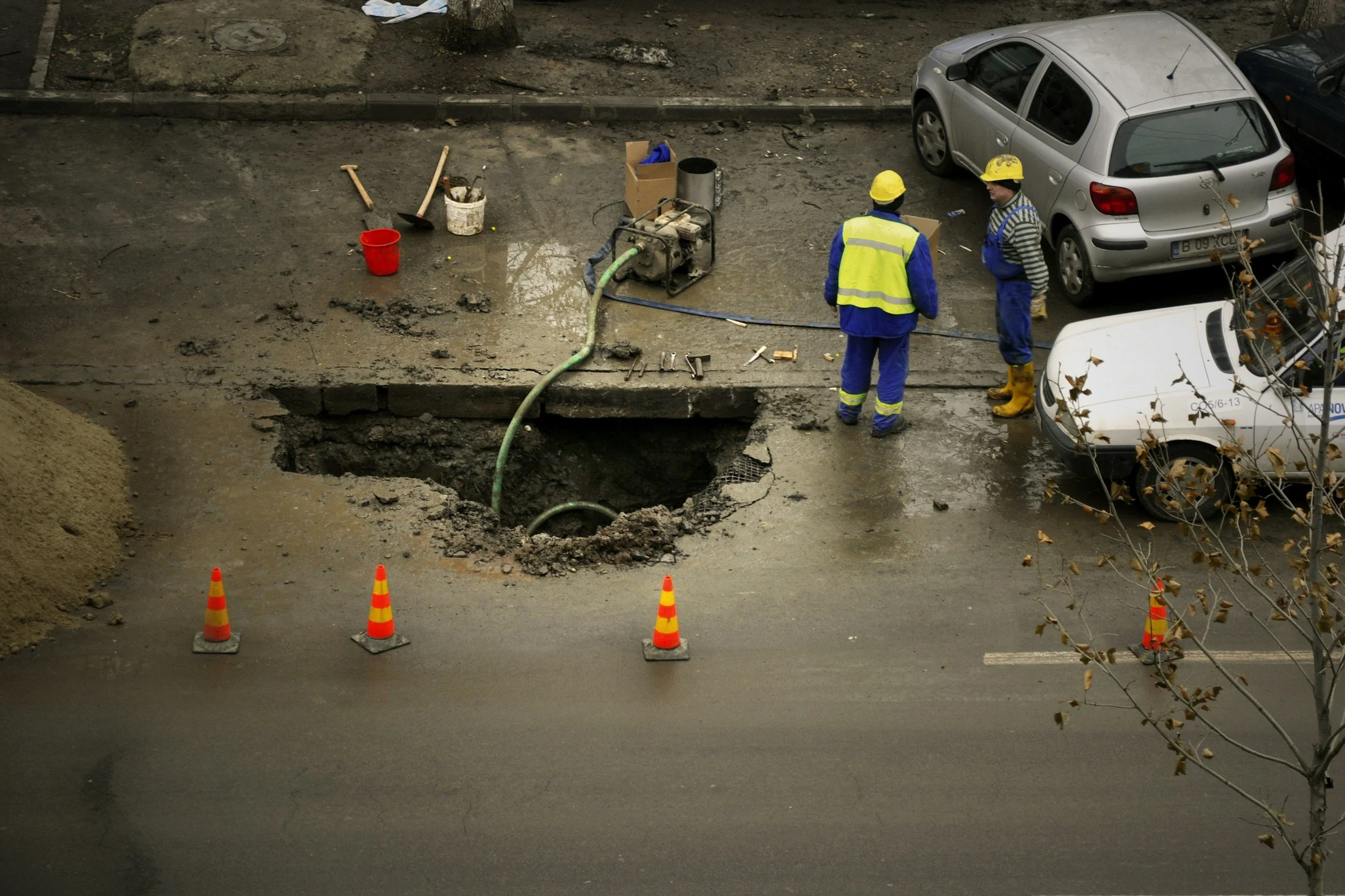 Construction workers in yellow safety helmets and blue uniforms working on a large hole in the road, with construction tools and equipment nearby, orange traffic cones blocking part of the road, and parked cars in the background.