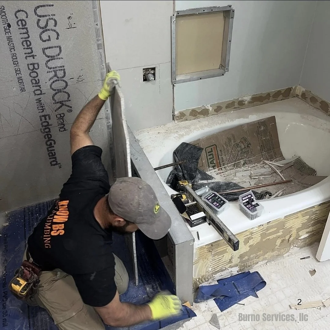 An elderly man with a shaved head wearing a black t-shirt with the word 'KNOW?' in orange text. He is working on a project, wearing a white glove on one hand, in a room with a brick wall and a window in the background.