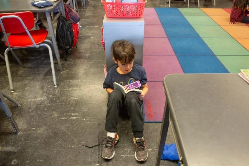 A young boy sitting on the floor in a classroom, reading a book. There are colorful mats, desks, and chairs around him.