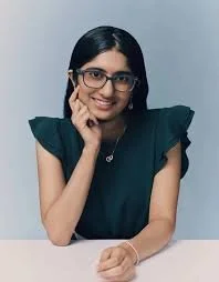 Young woman with black hair and glasses smiling, wearing a dark green top, resting her chin on her hand.