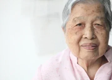 Close-up of an elderly Asian woman with gray hair smiling, wearing a pink shirt.