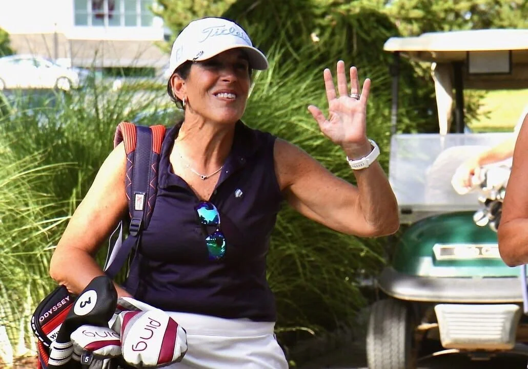 A woman in a golf outfit, wearing a white cap, black sleeveless top, and white shorts, smiling and waving. She has a golf club with a glove attached and sunglasses hanging from her top. Background includes tall green plants and part of a golf cart.