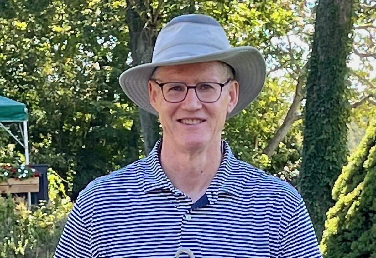 A smiling man wearing glasses, a wide-brimmed hat, and a striped polo shirt, standing outdoors in front of trees and greenery.