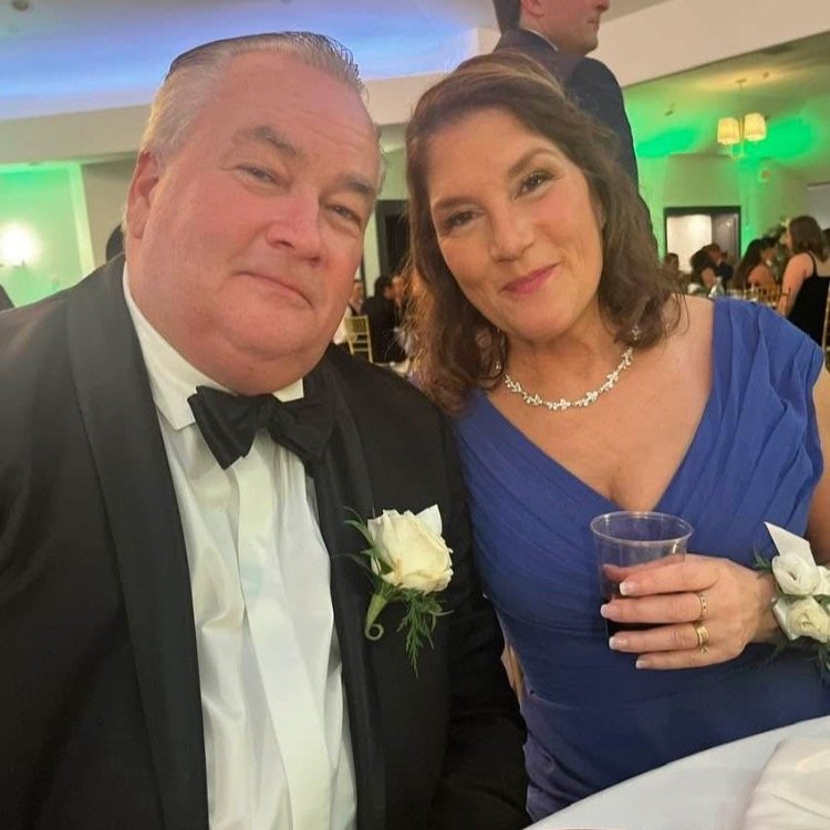 A man in a tuxedo and a woman in a blue dress sitting at a wedding reception, smiling at the camera. The man has a white boutonniere, and the woman has a corsage and is holding a drink.