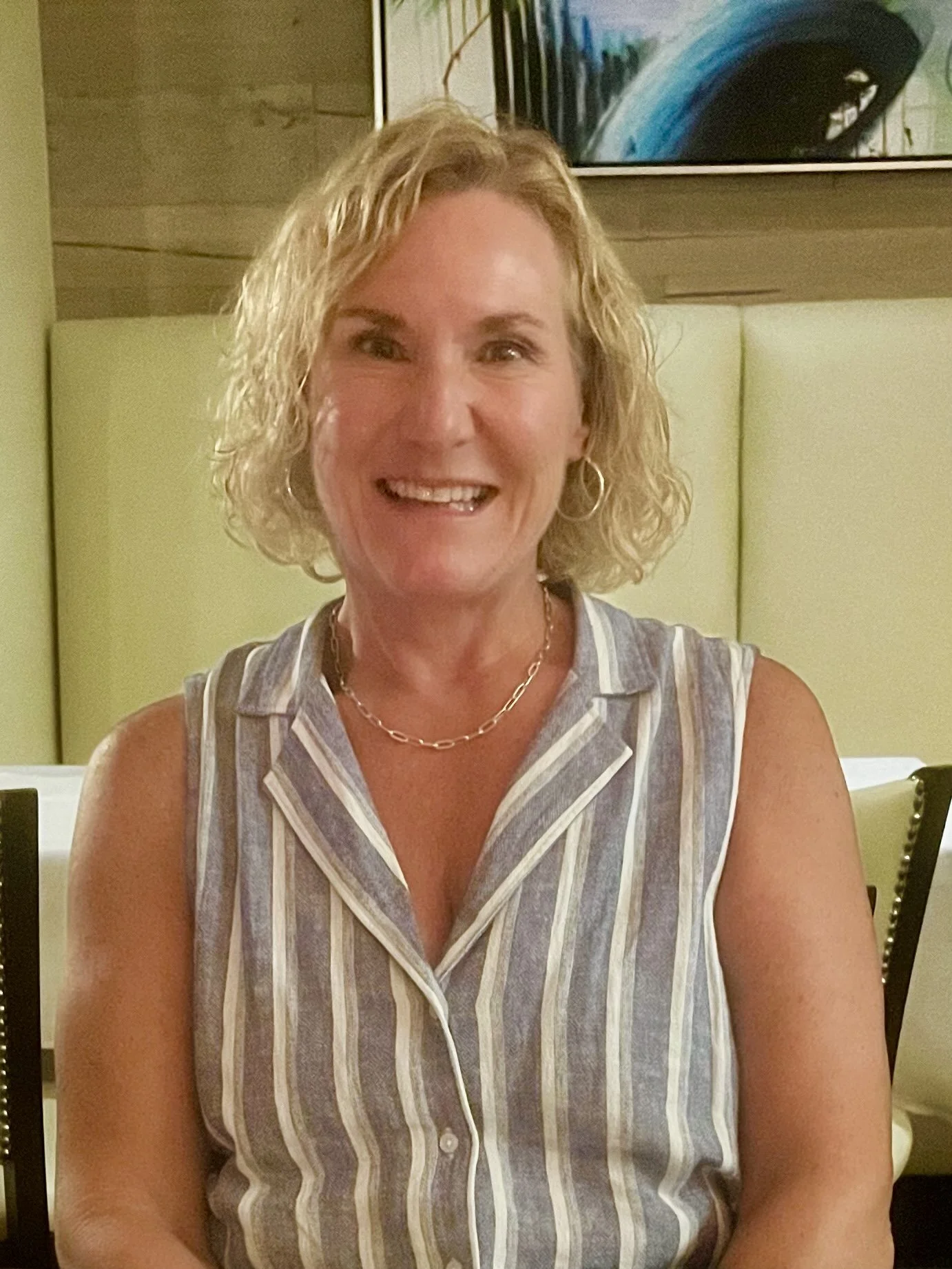 A woman with curly blonde hair smiling, wearing a sleeveless striped shirt and gold jewelry, sitting in a restaurant with a framed abstract painting on the wall behind her.