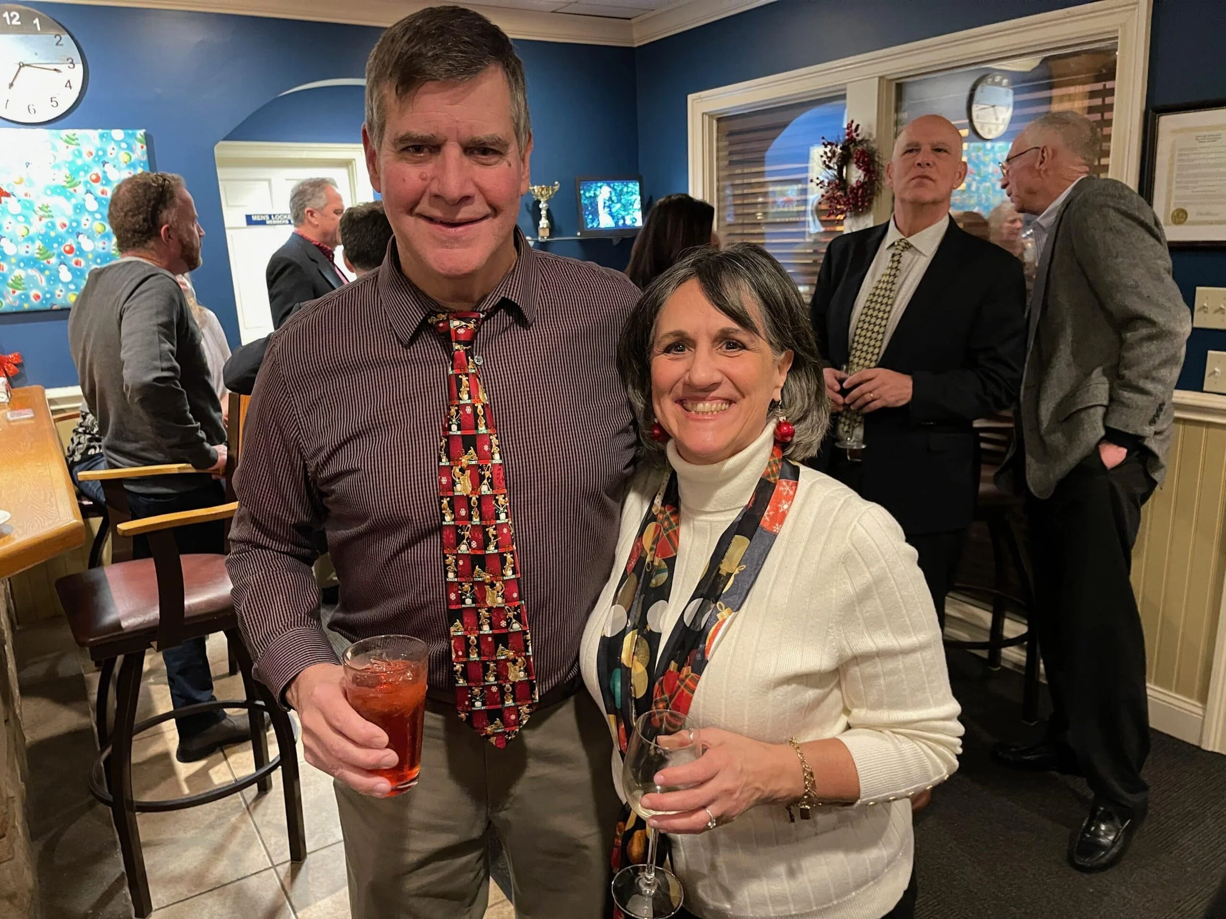 A man and woman smiling at a holiday party, each holding a drink, with other guests talking in the background.