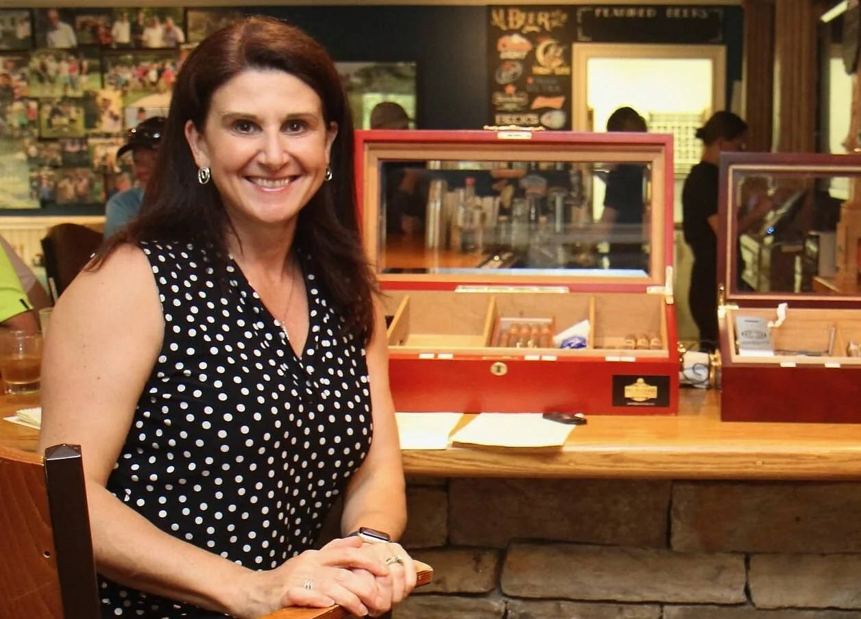 A woman with long dark hair, wearing a black sleeveless polka dot dress, smiling and sitting at a bar counter with cigar humidors behind her in a cigar lounge or smoke shop.