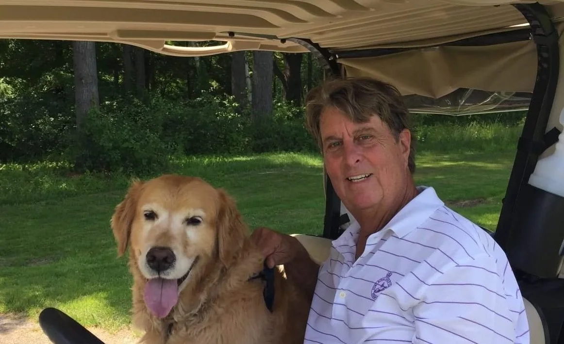 A man with brown hair smiling at the camera, sitting next to a golden retriever with its tongue out, in a golf cart outdoors with green grass and trees in the background.