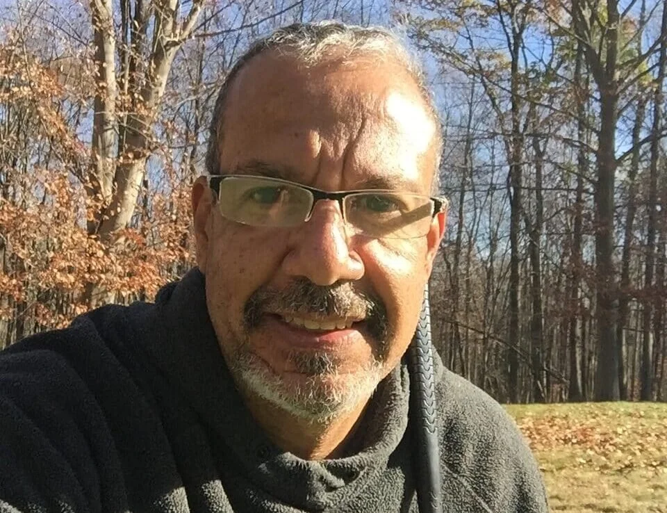 Close-up of a middle-aged man with glasses, a beard, and a mustache outdoors in a wooded area, smiling at the camera.