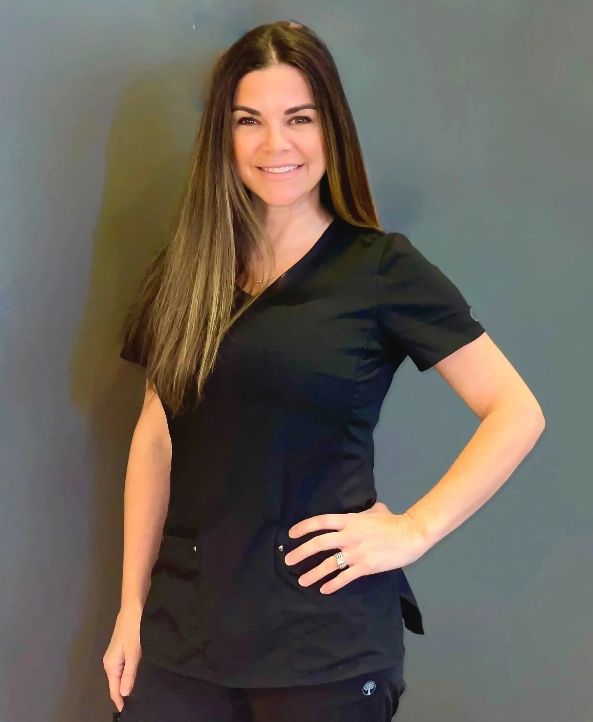 A woman with long brown hair wearing black medical scrubs, standing against a gray background, smiling with her left hand on her hip.
