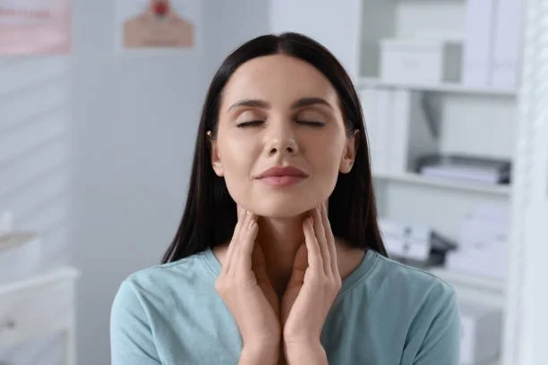 Woman with dark hair gently touching her throat with both hands, eyes closed, appears to be experiencing throat discomfort or pain.