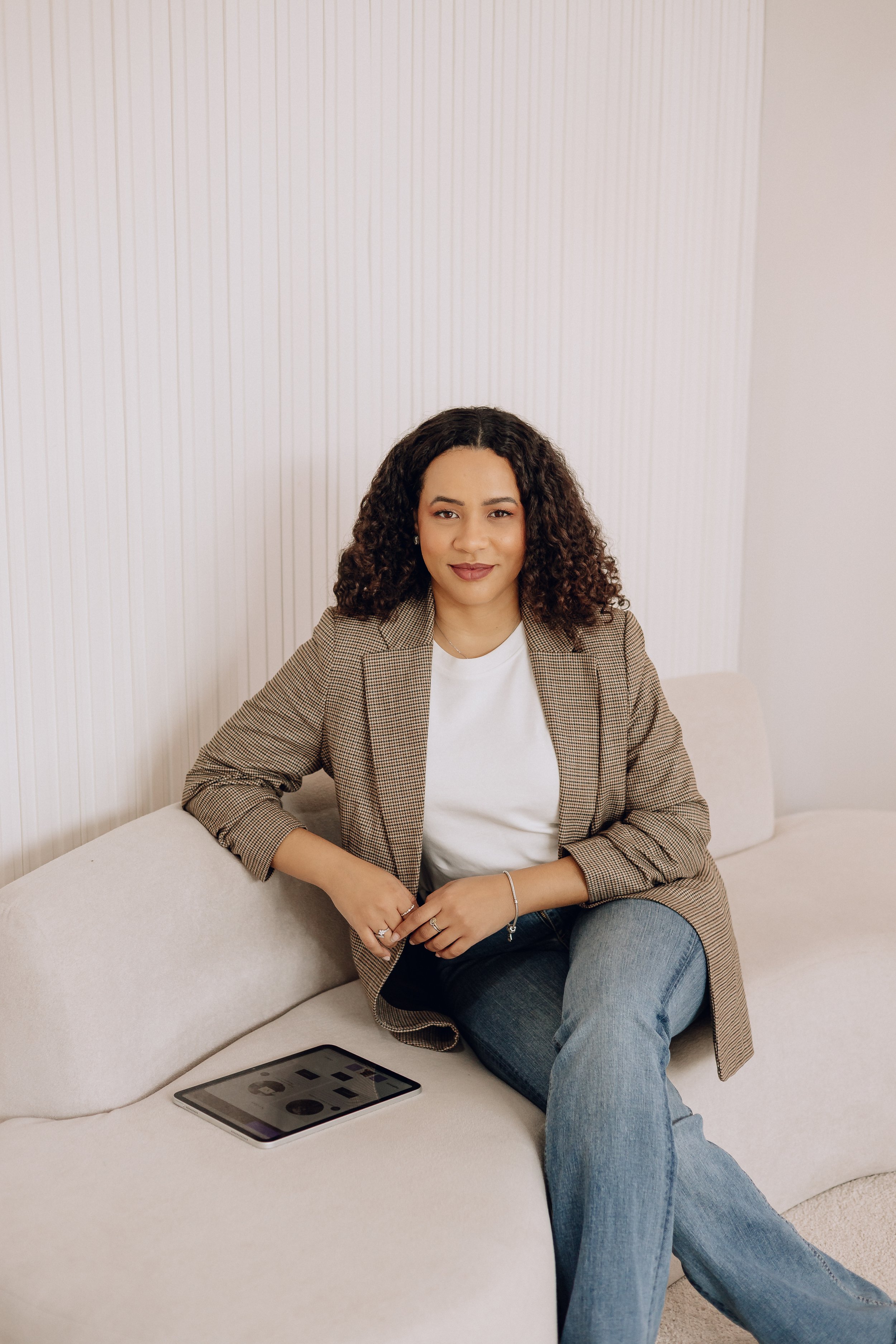 A woman with curly hair sitting on a light-colored sofa with an iPad on her lap, wearing a blazer, white t-shirt, and jeans in a minimalist room.