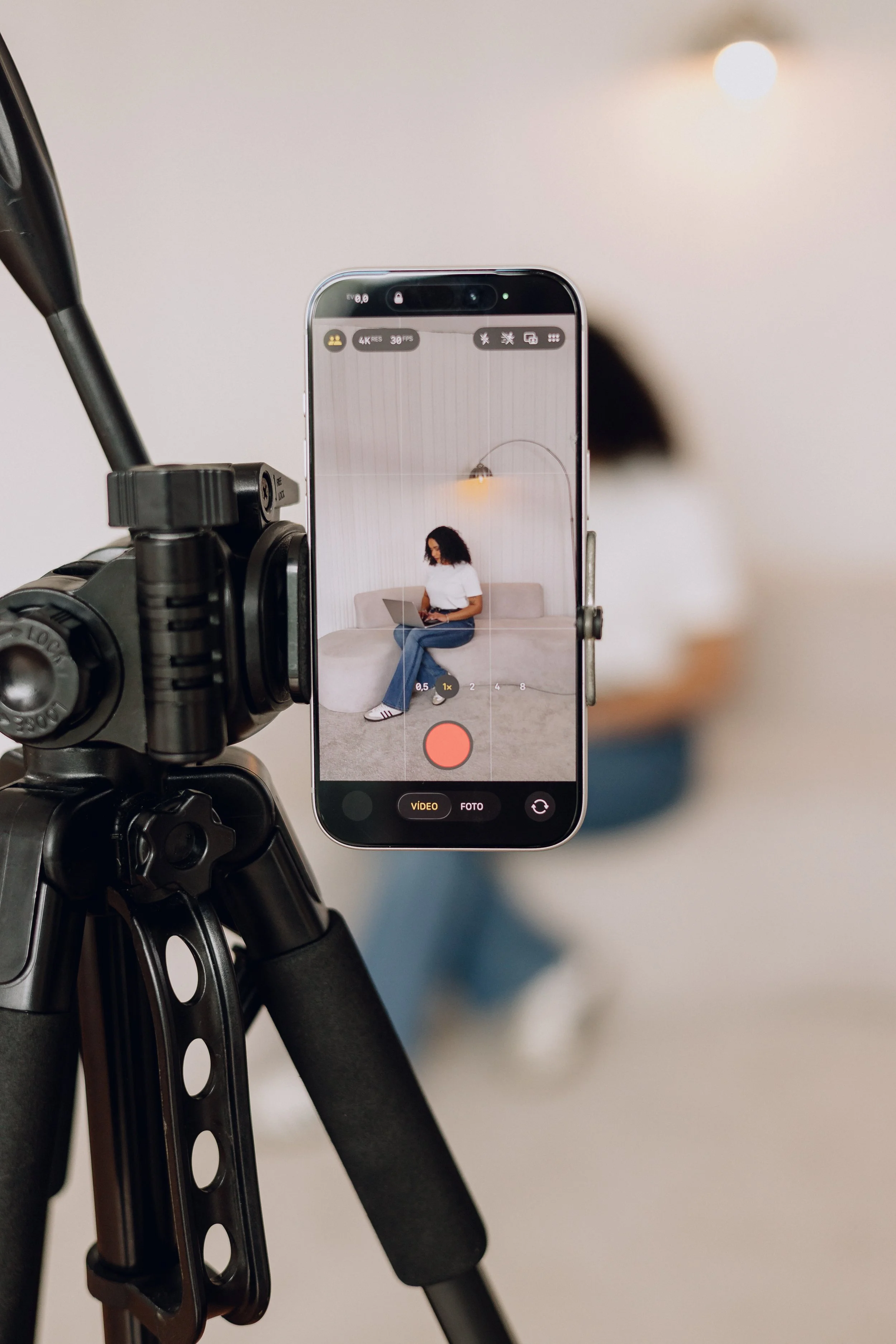 A woman sitting on a beige couch with a laptop, seen through a camera phone mounted on a tripod.