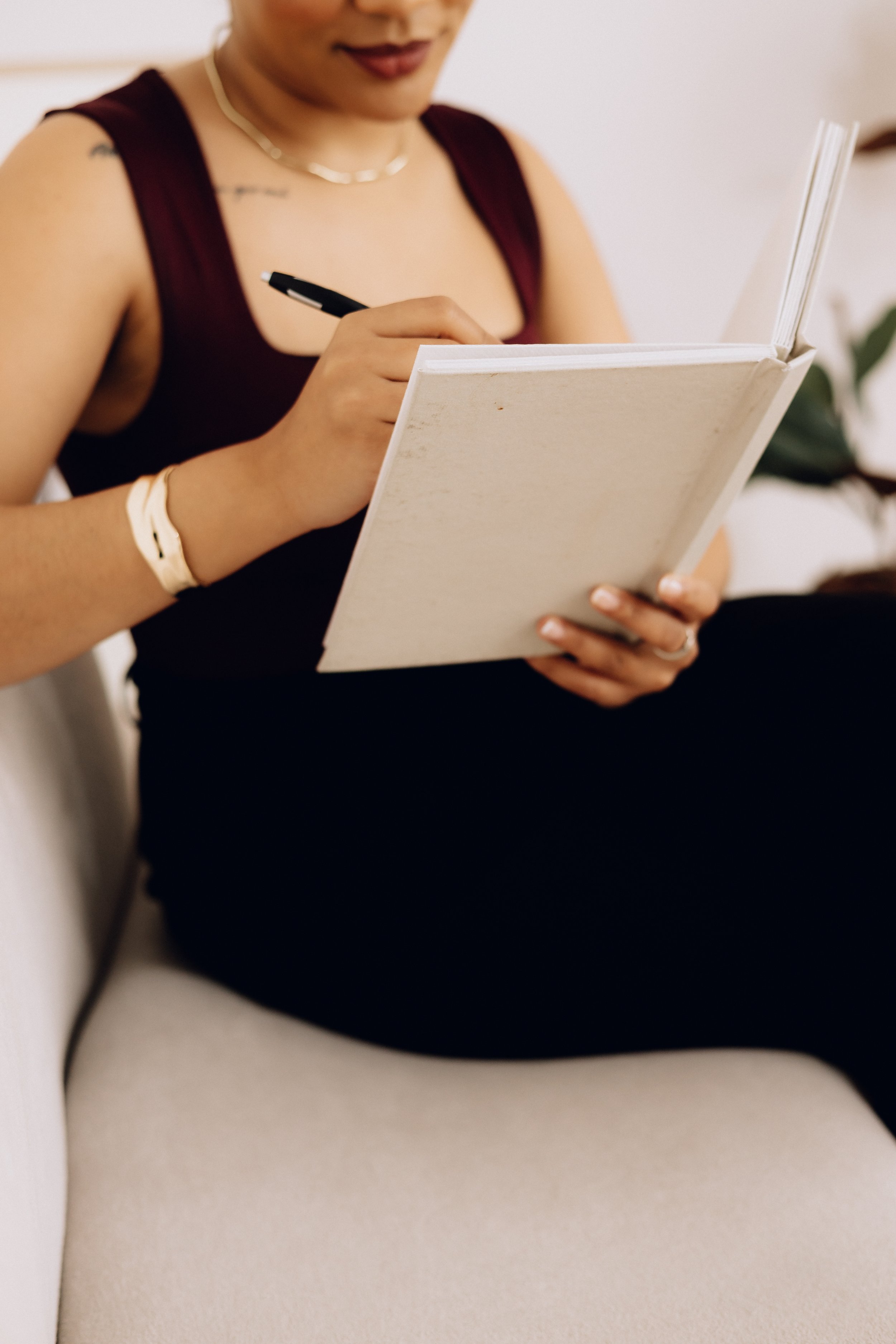 A woman sitting on a white couch, writing in a notebook with a black pen.