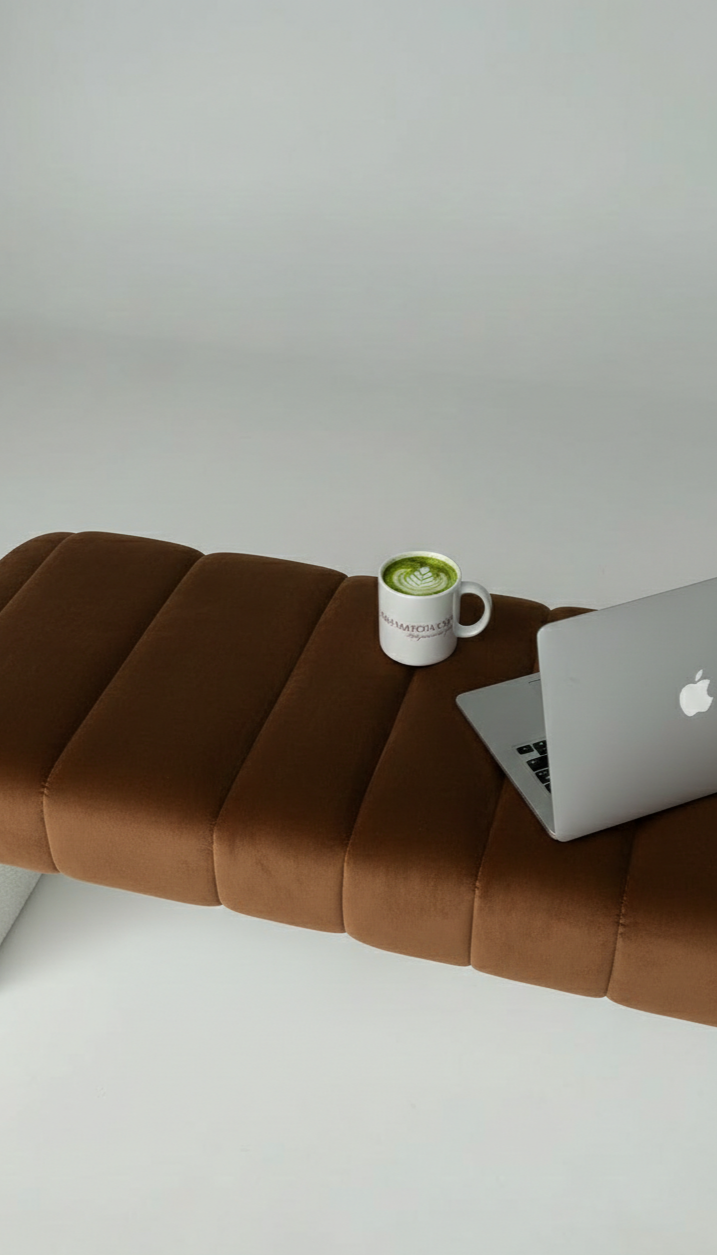A brown cushioned bench with a cup of matcha latte and a silver Apple MacBook on it, against a plain white wall.