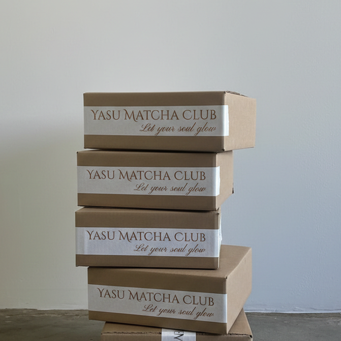 Four cardboard boxes stacked vertically against a plain wall, labeled 'YASU MATCHA CLUB' with the slogan 'Let your soul glow' on each.