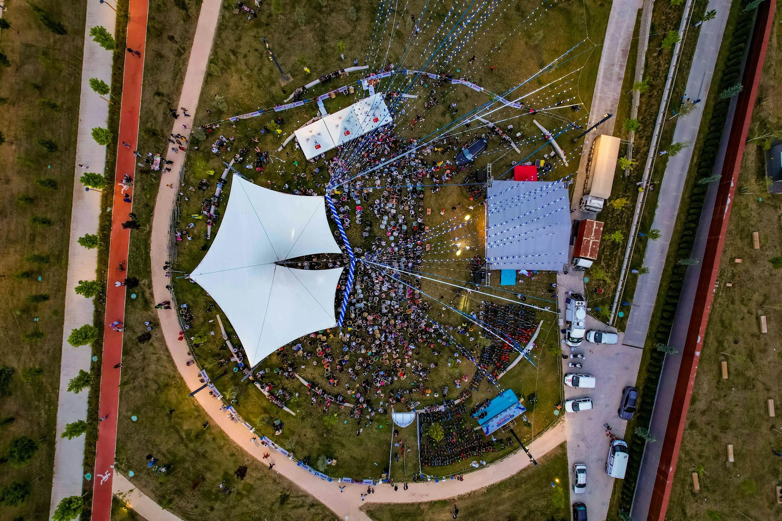 An aerial view of a crowded outdoor event with two large white tents, a stage, and various smaller tents, surrounded by a grassy area, walking paths, and parked cars.