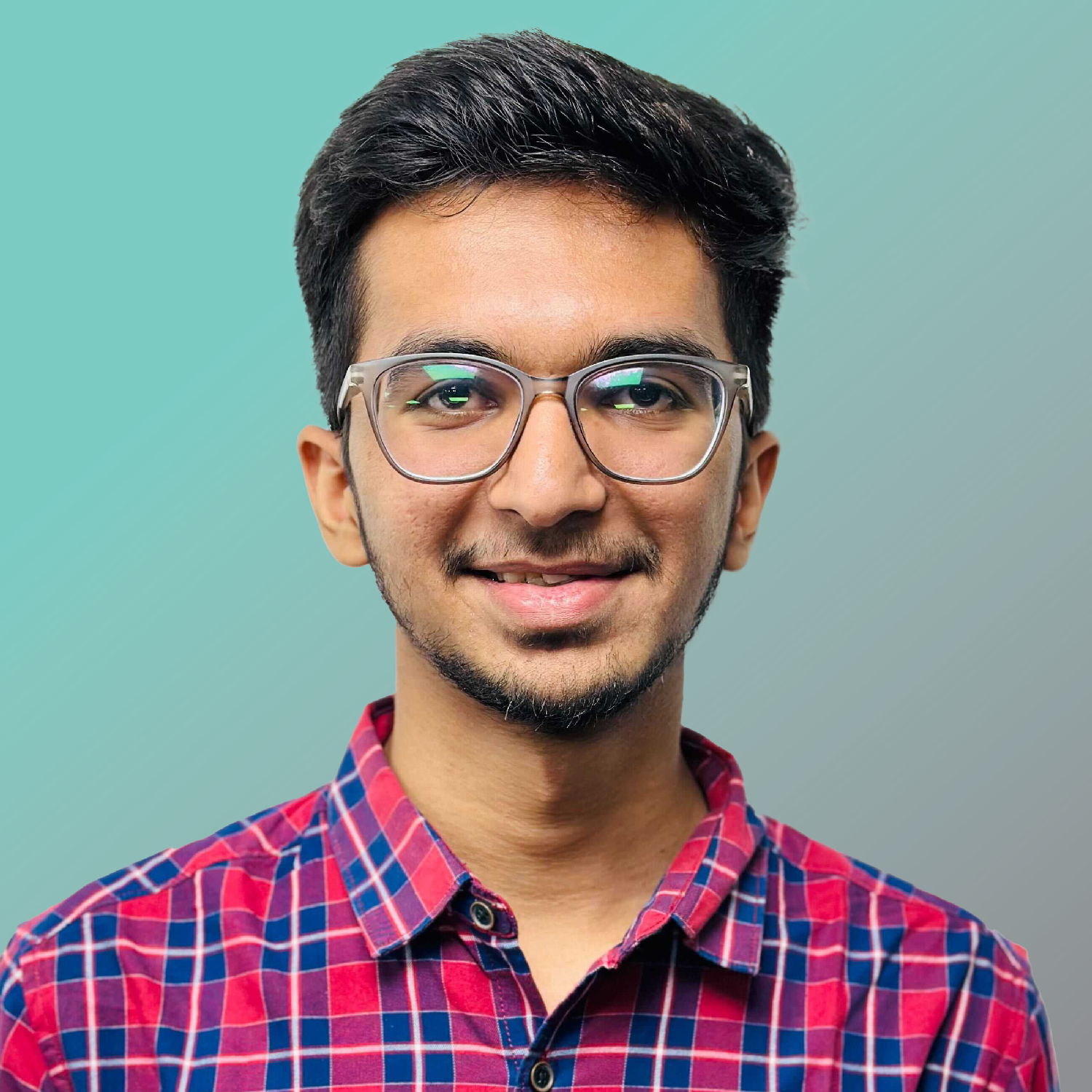 A young man with short dark hair, glasses, and a trimmed beard, wearing a red and blue checked shirt, smiling at the camera against a light blue background.