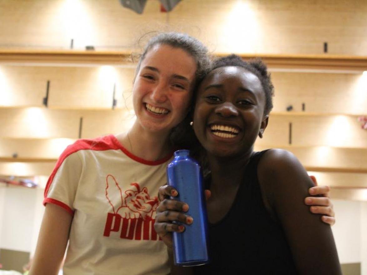 Twee jonge vrouwen die lachen, een van hen houdt een blauwe waterfles vast, in een houten sporthal of zaal.
