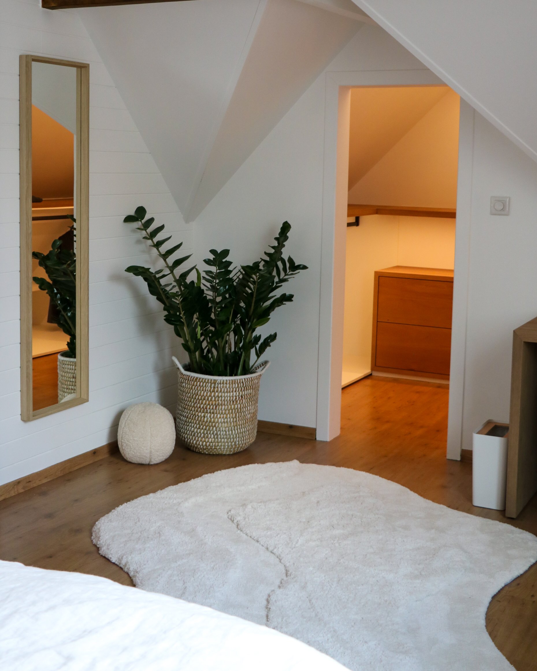 Een modern ingerichte slaapkamer met een grote plant in een mand, een staande spiegel, een zacht wit tapijt met organische vorm op een houten vloer en toegang tot de op maat gemaakte inloopkast onder schuin dak.