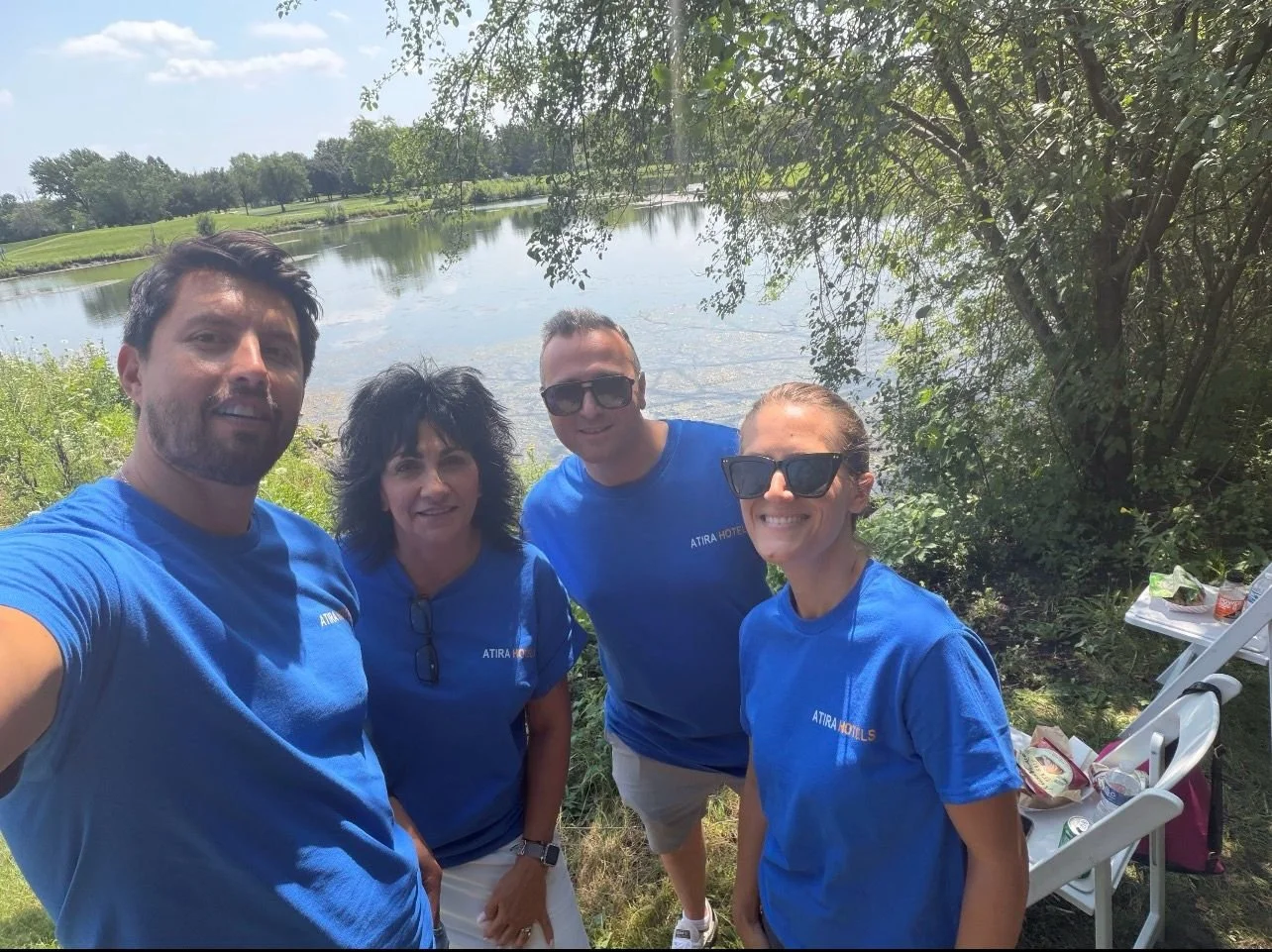 Four people standing outdoors by a pond, smiling at the camera, wearing blue shirts, with trees and a table with food in the background.