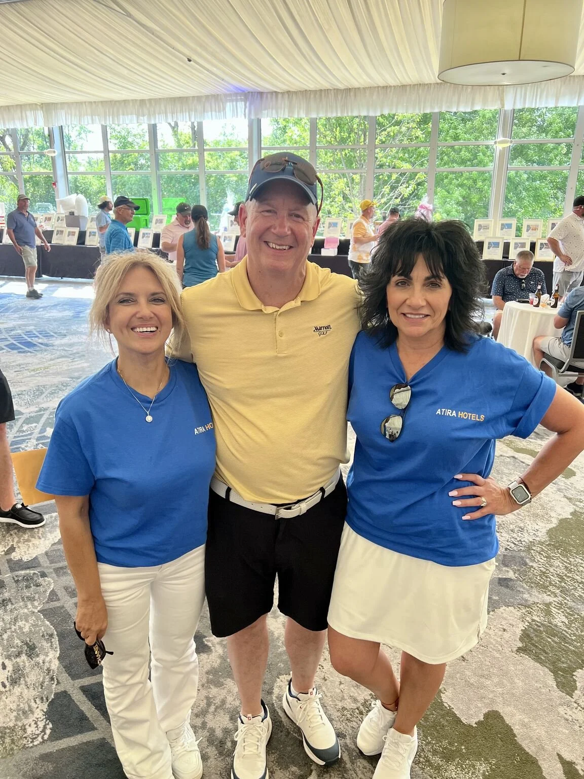 Three people standing indoors at a social event, smiling at the camera. Two women are wearing blue ATIRA HOTELS shirts, and a man in between them is dressed in a yellow polo shirt and black shorts. The background features large windows with a view of green trees and people browsing displayed items.