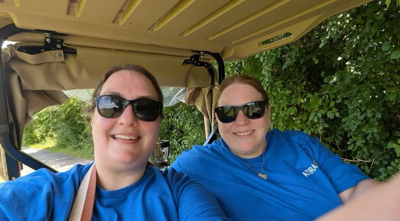 Two women wearing blue shirts and sunglasses sitting inside a golf cart, smiling for a selfie, with green bushes and a tree-lined road in the background.