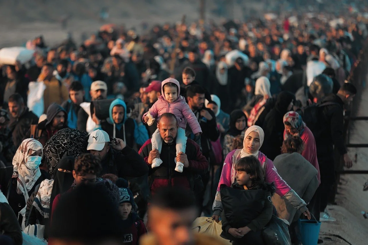 Une grande foule de personnes, dont beaucoup portent des vêtements chauds, se rassemble probablement lors d'une migration ou d'une crise humanitaire, avec quelques femmes portant des foulards et des enfants sur les épaules.