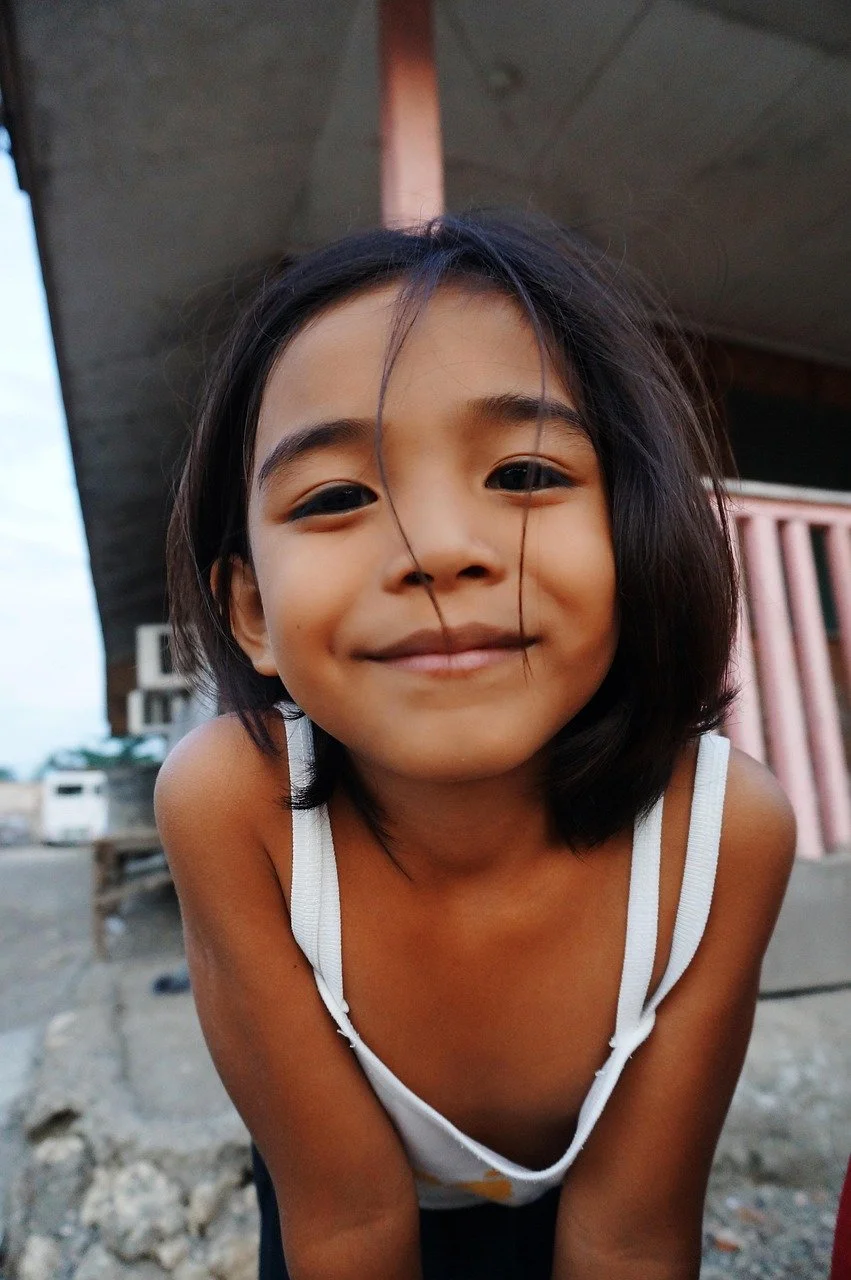 Jeune fille souriante avec cheveux noirs, portant un haut blanc à fines bretelles, en extérieur, arrière-plan d'une structure en béton et bois.