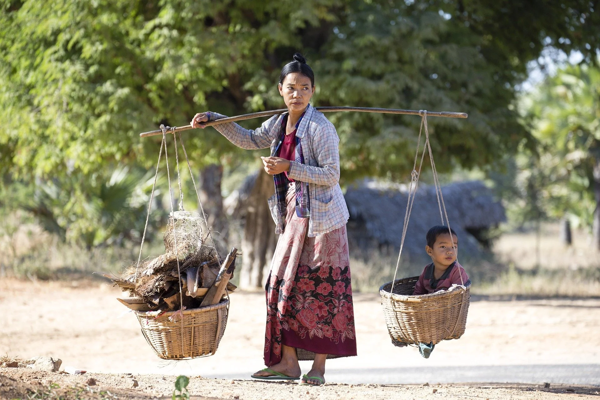 Une femme portant des paniers suspendus d'une bambou sur une sorcière, avec un enfant dans un panier, dans un environnement rural avec des arbres verts.