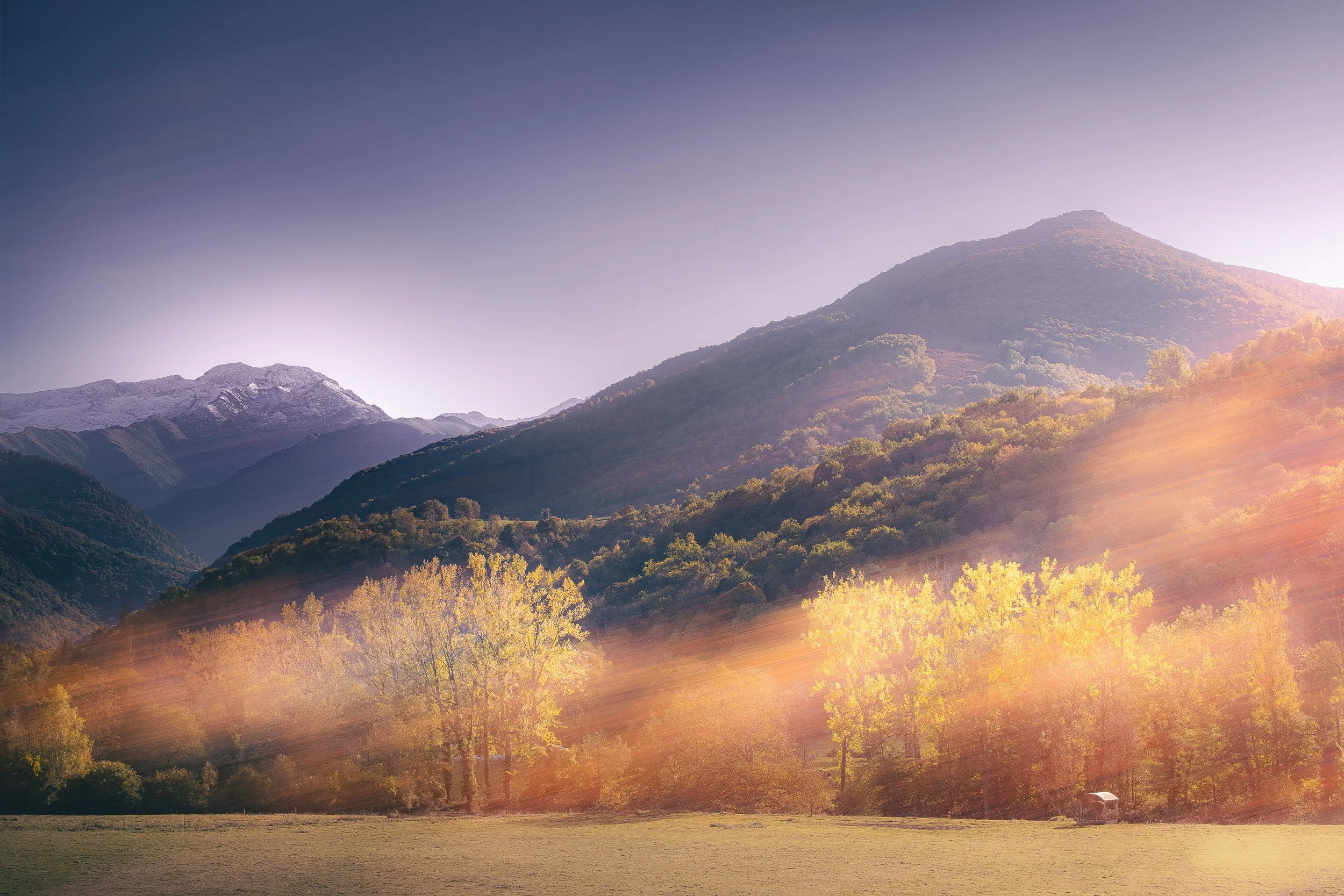 Paysage de montagnes avec des sommets enneigés, des collines verdoyantes, arbres colorés en automne, et rayons de soleil à l'aube ou au crépuscule.
