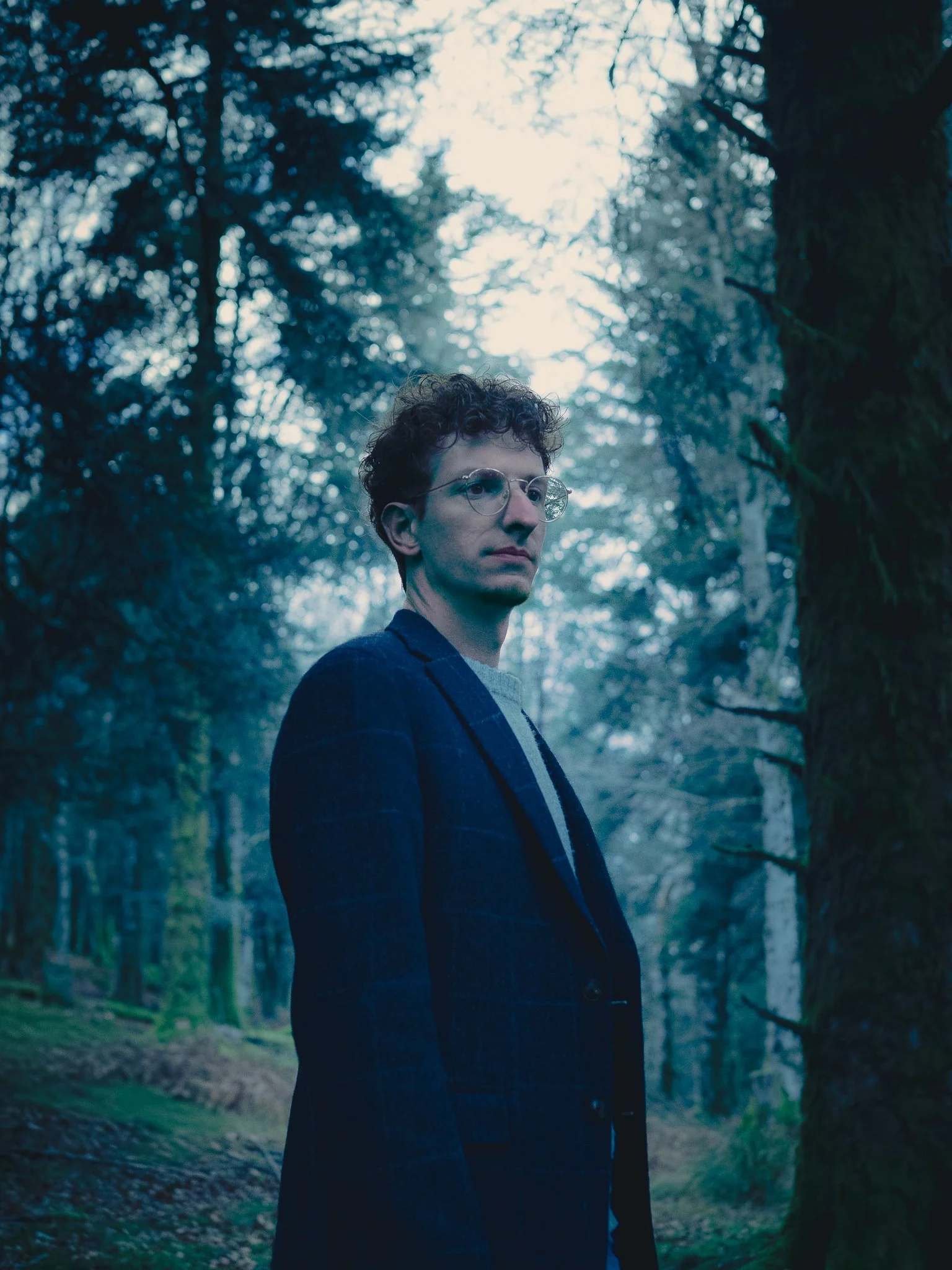 Portrait de Damien Trussardi. Description : jeune homme avec lunettes, portant un blazer foncé, dans une forêt dense.