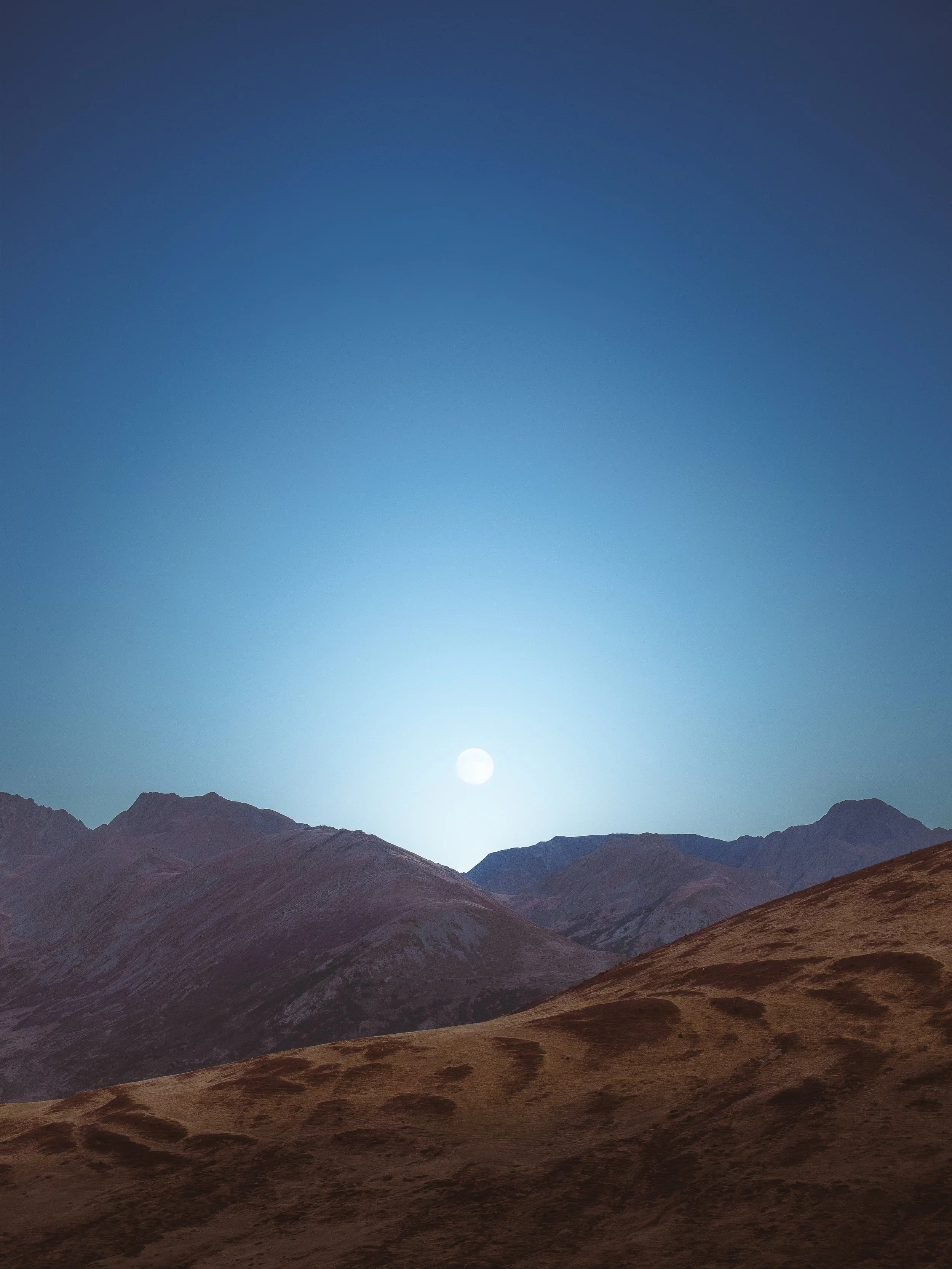 Paysage avec des montagnes sous un ciel clair, avec la lune visible à l'horizon.