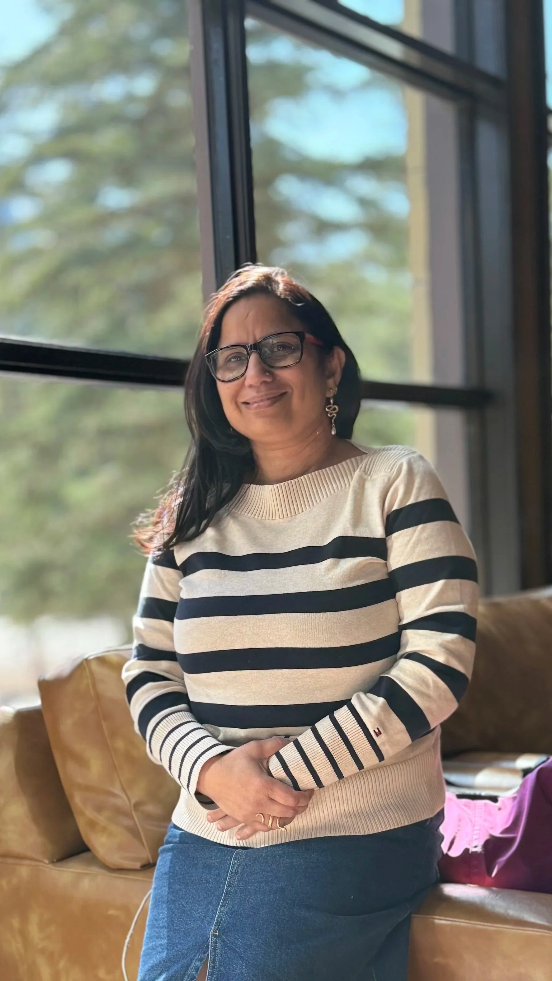 A woman with glasses and earrings smiling, wearing a striped sweater and blue jeans, sitting in front of a window with trees outside.