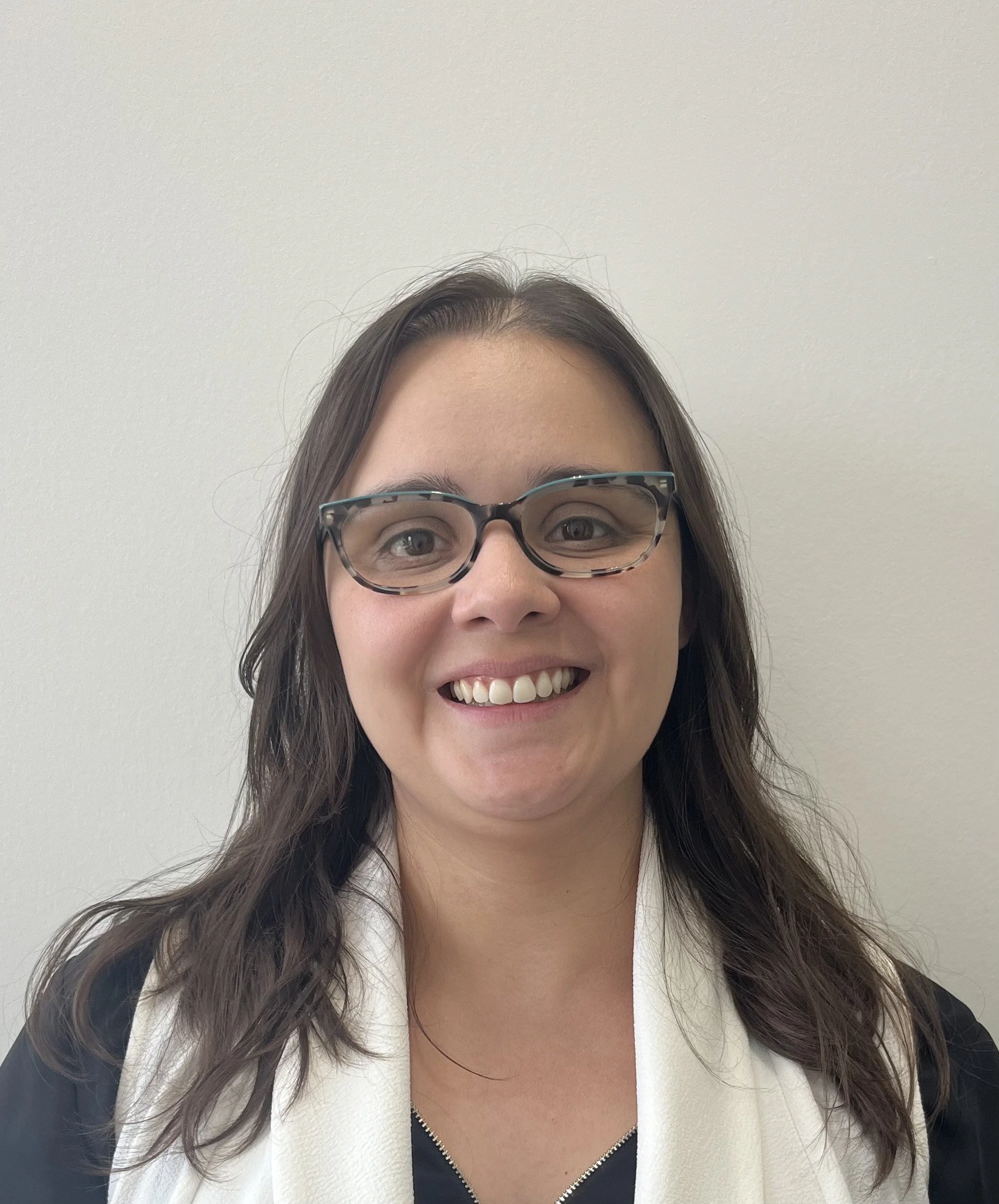 A woman with long dark hair, glasses, and a white scarf smiling in front of a plain white wall.