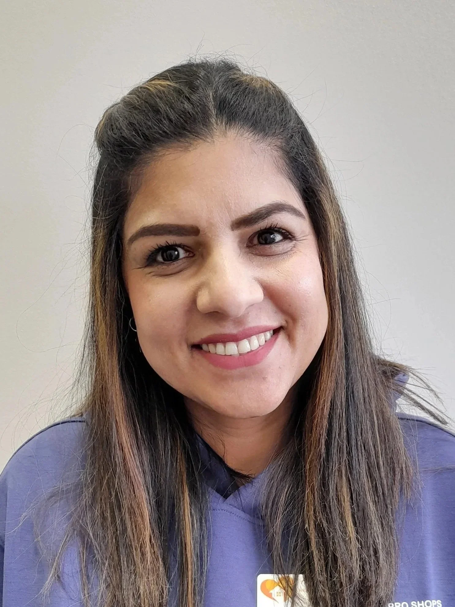 Close-up of a smiling woman with long brown hair, wearing a purple top, against a plain light-colored background.