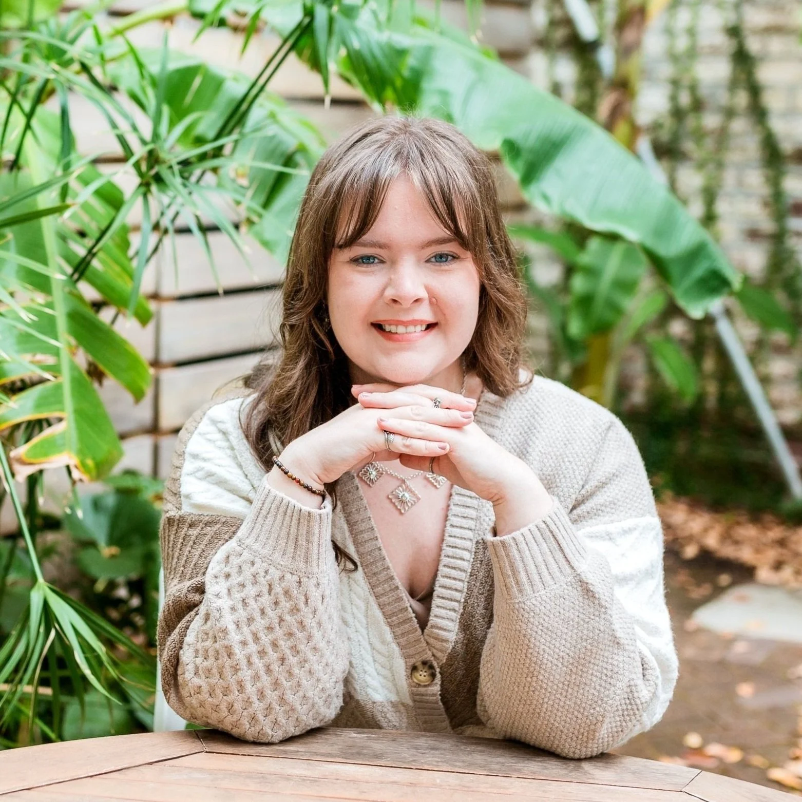Meghan Lovell, a labor and postpartum doula, smiling, with brown hair and blue eyes, sitting outdoors at a wooden table surrounded by large green leafy plants.