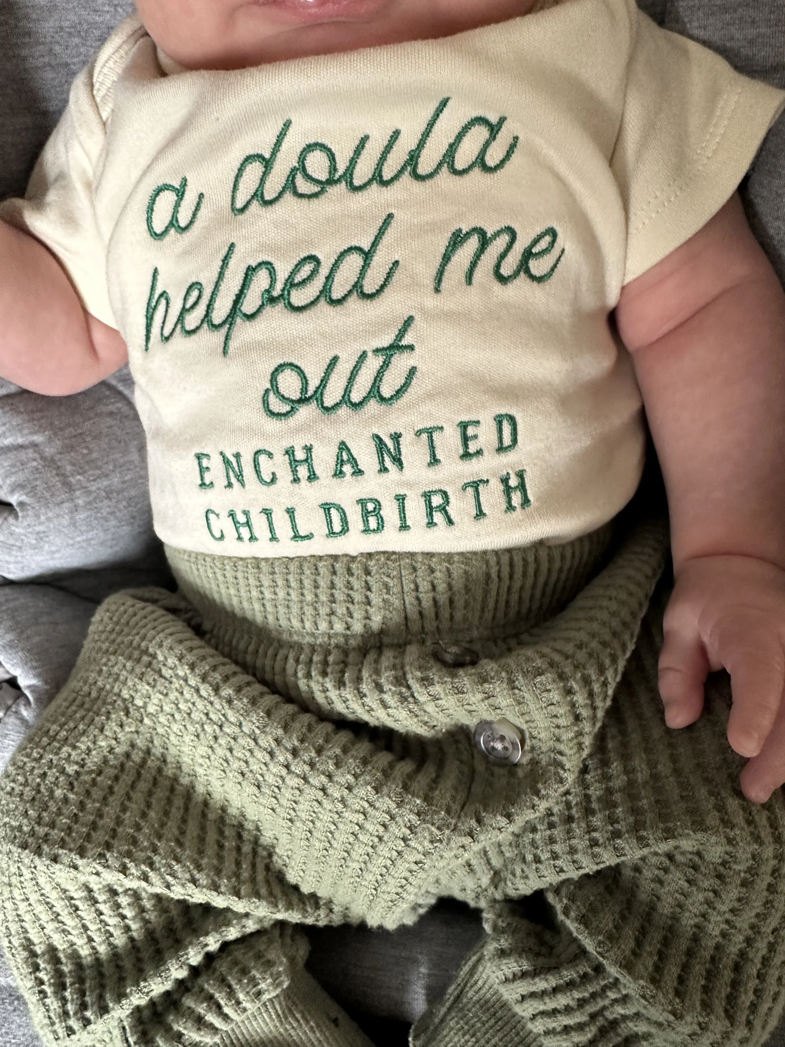 Close-up of a baby wearing a cream-colored shirt with green embroidery that reads: 'a double helped me out, enchanted childhood.' The baby is being held gently, with part of a gray sleeve visible in the background and a hoodie with a textured knit pattern in the lower part of the image.