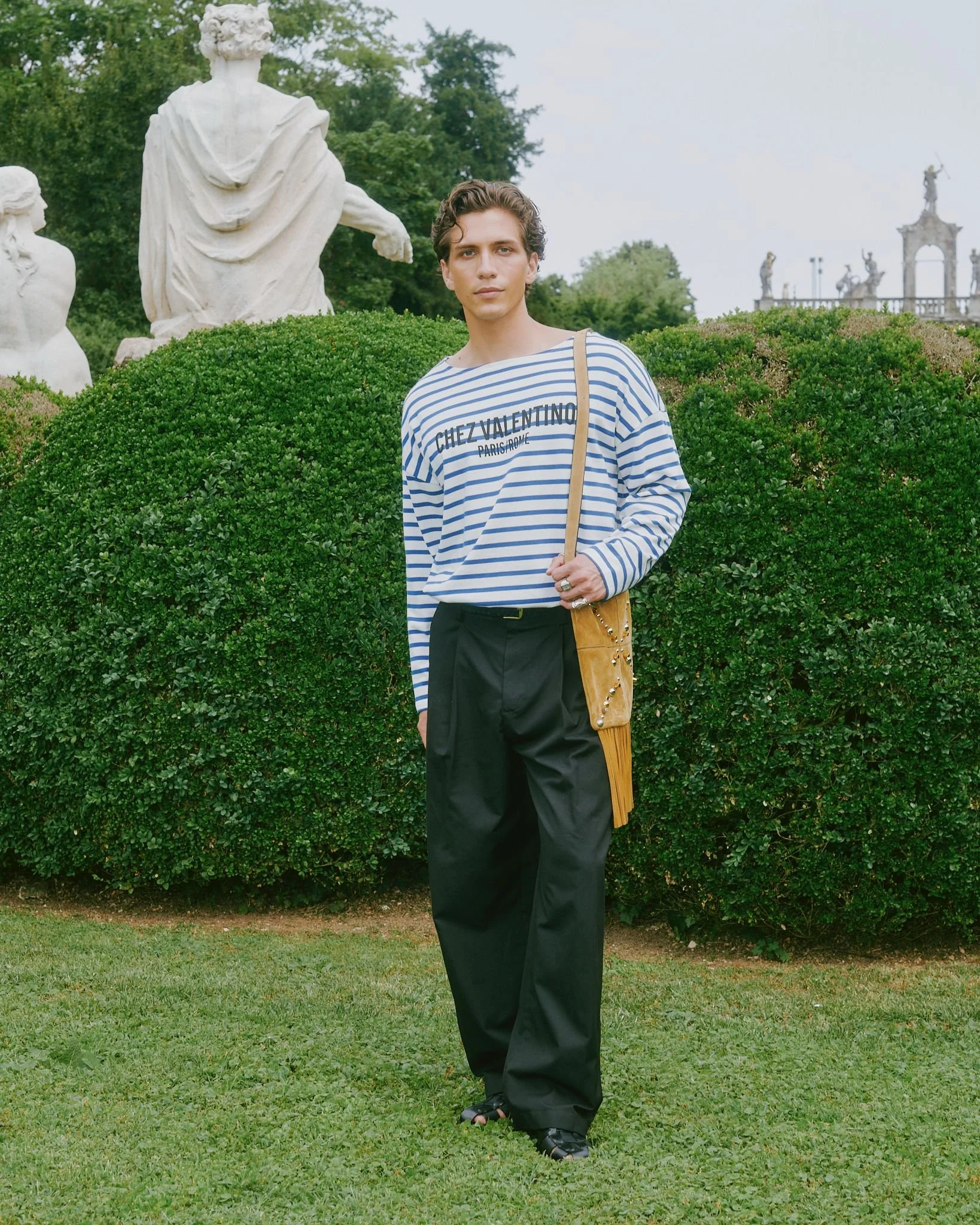 A person with wavy short hair standing outdoors on grass in front of a large bush, wearing a striped shirt, black pants, and sandals, carrying a yellow shoulder bag, with classical sculptures in the background.