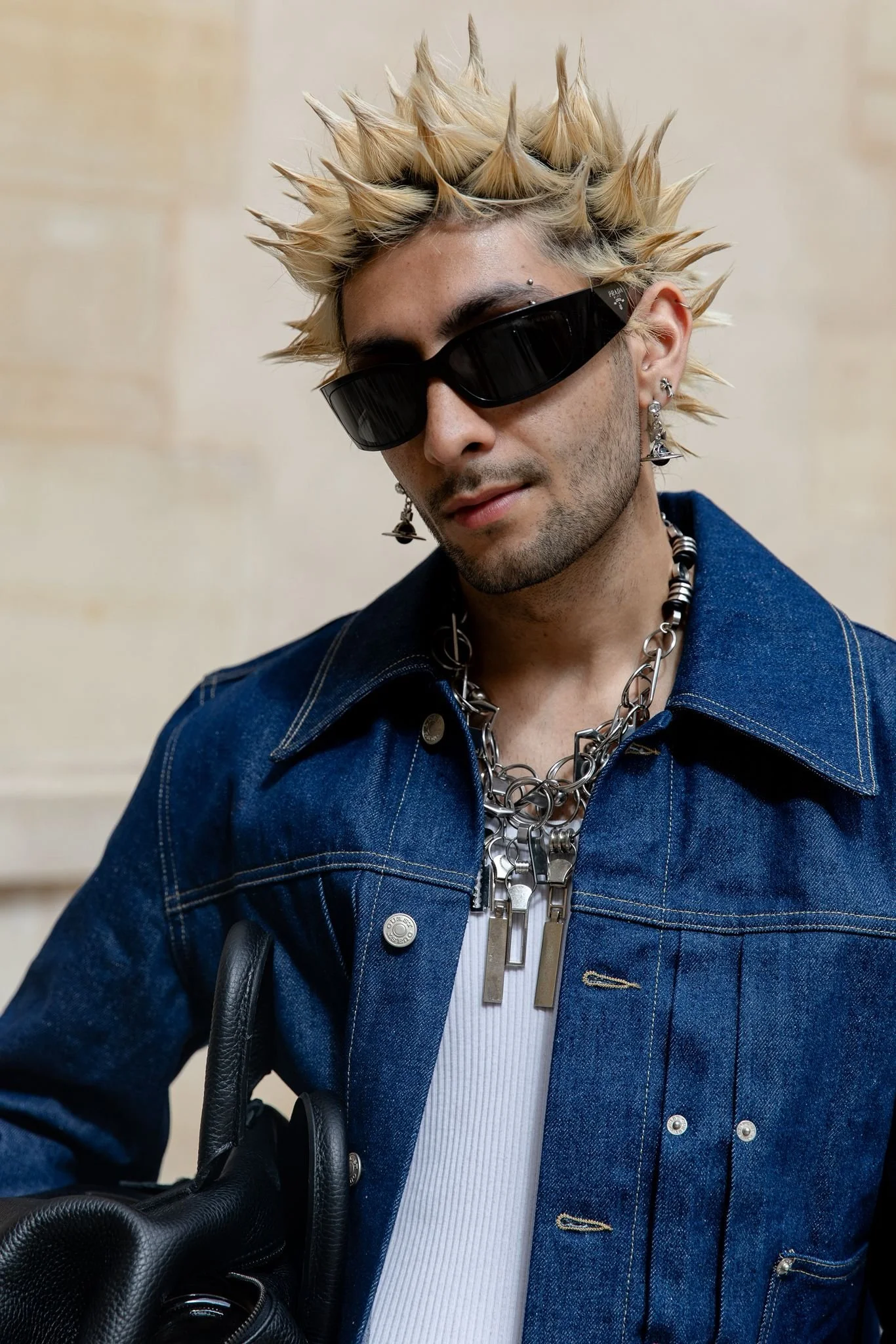 A person with spiked blond hair wearing black sunglasses, a denim jacket, and chunky jewelry.