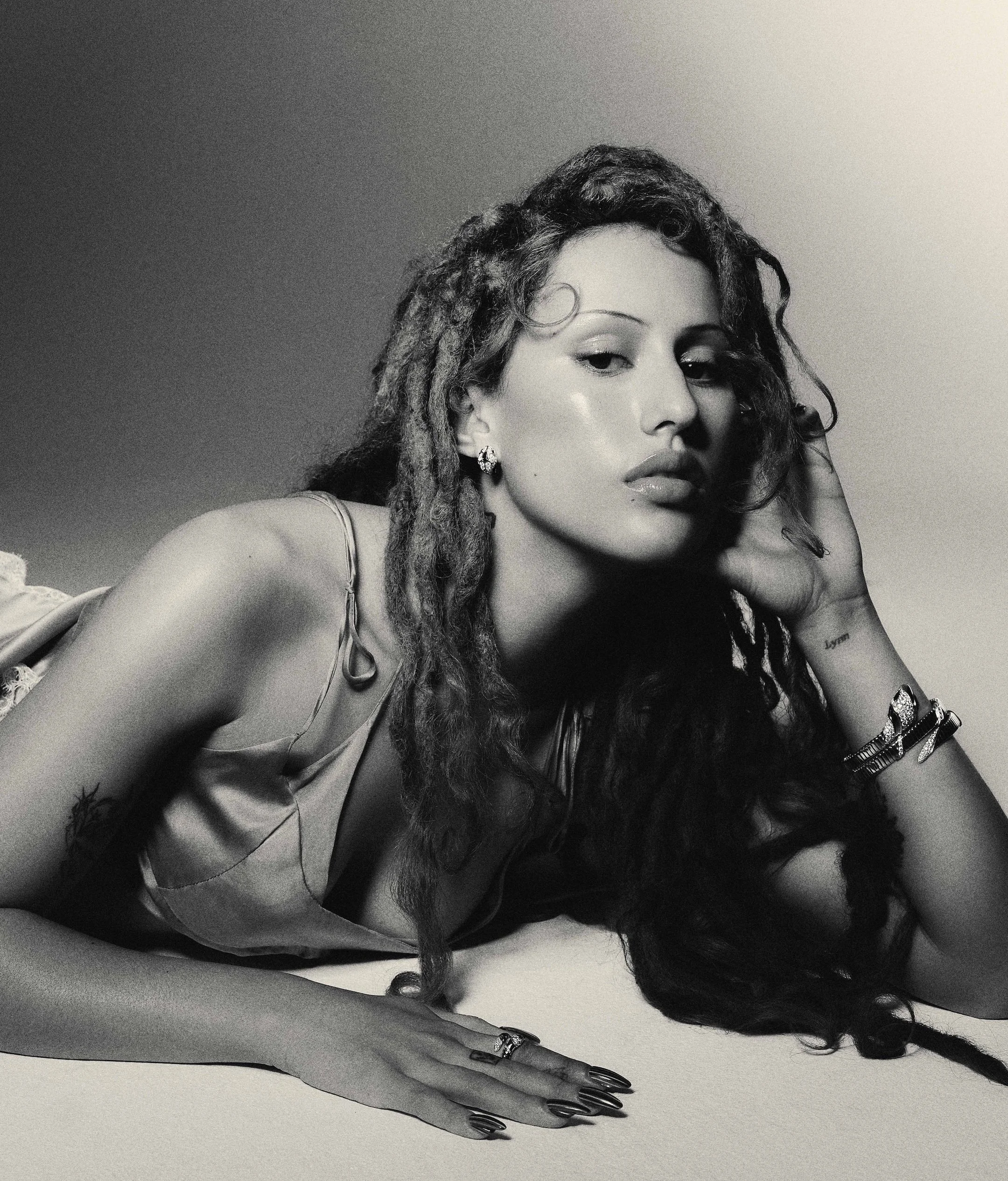 Person with long black hair styled in buns, wearing a black top and red fishnet tights with intricate nail designs, sitting against a blue wall.