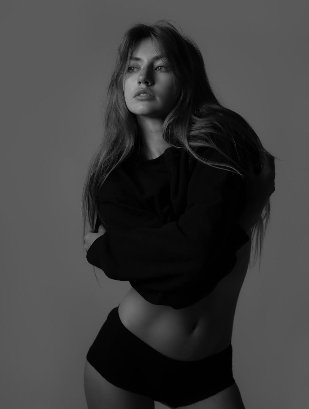 Black and white photo of a woman in a relaxed pose wearing a sweatshirt and underwear, with long flowing hair and thoughtful expression.