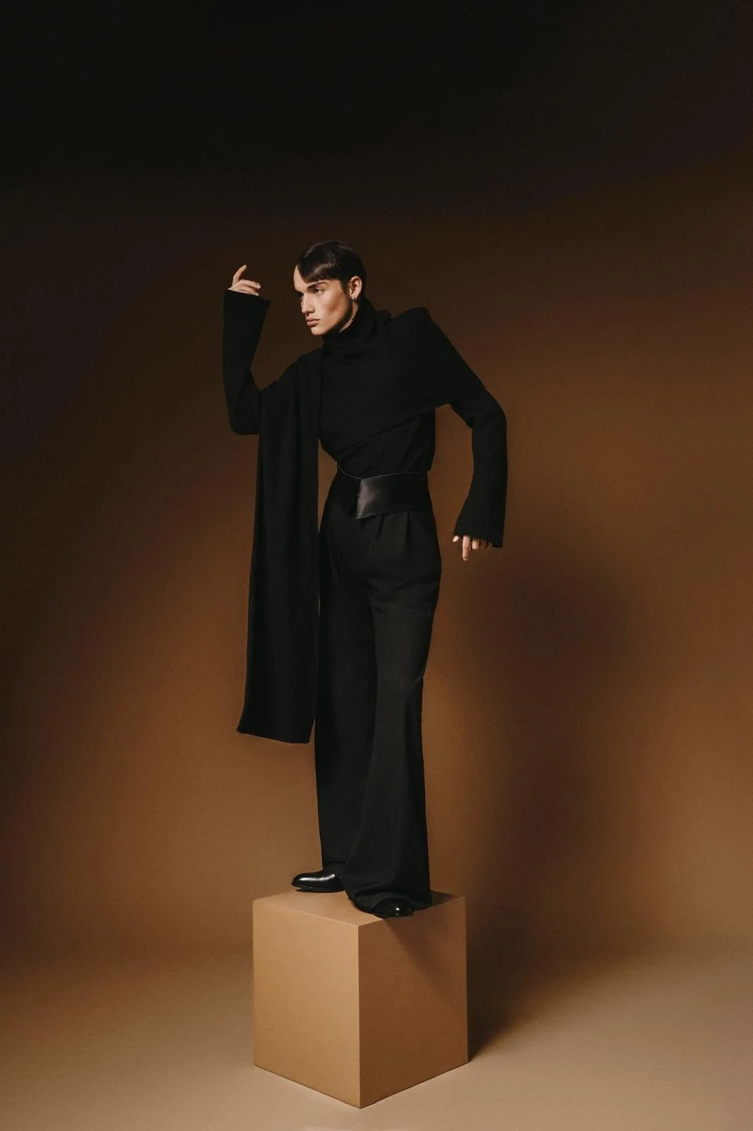 Person in black outfit standing on a brown box against a brown background