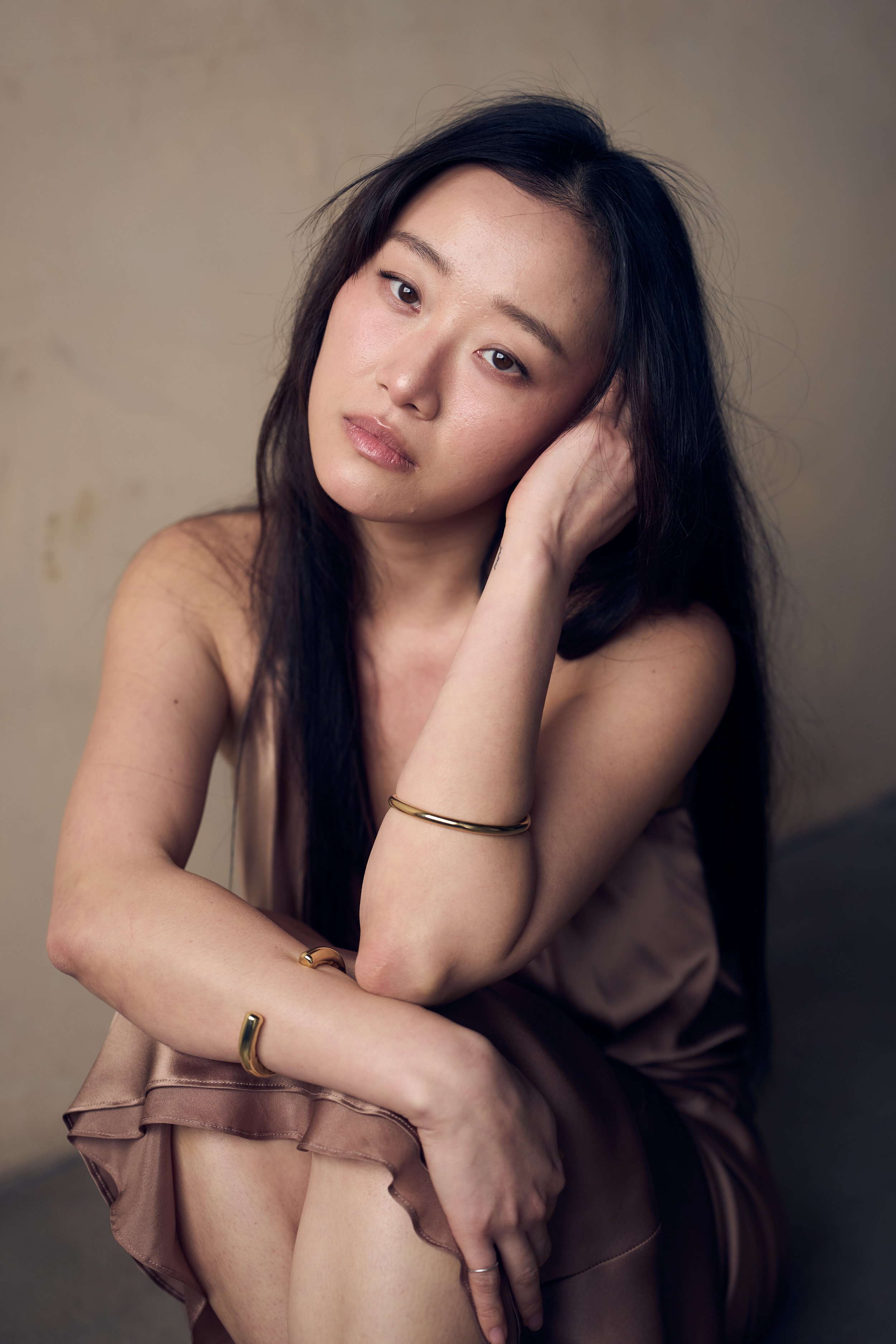 Woman with long hair sitting, wearing a brown dress and gold bracelets, looking at the camera.