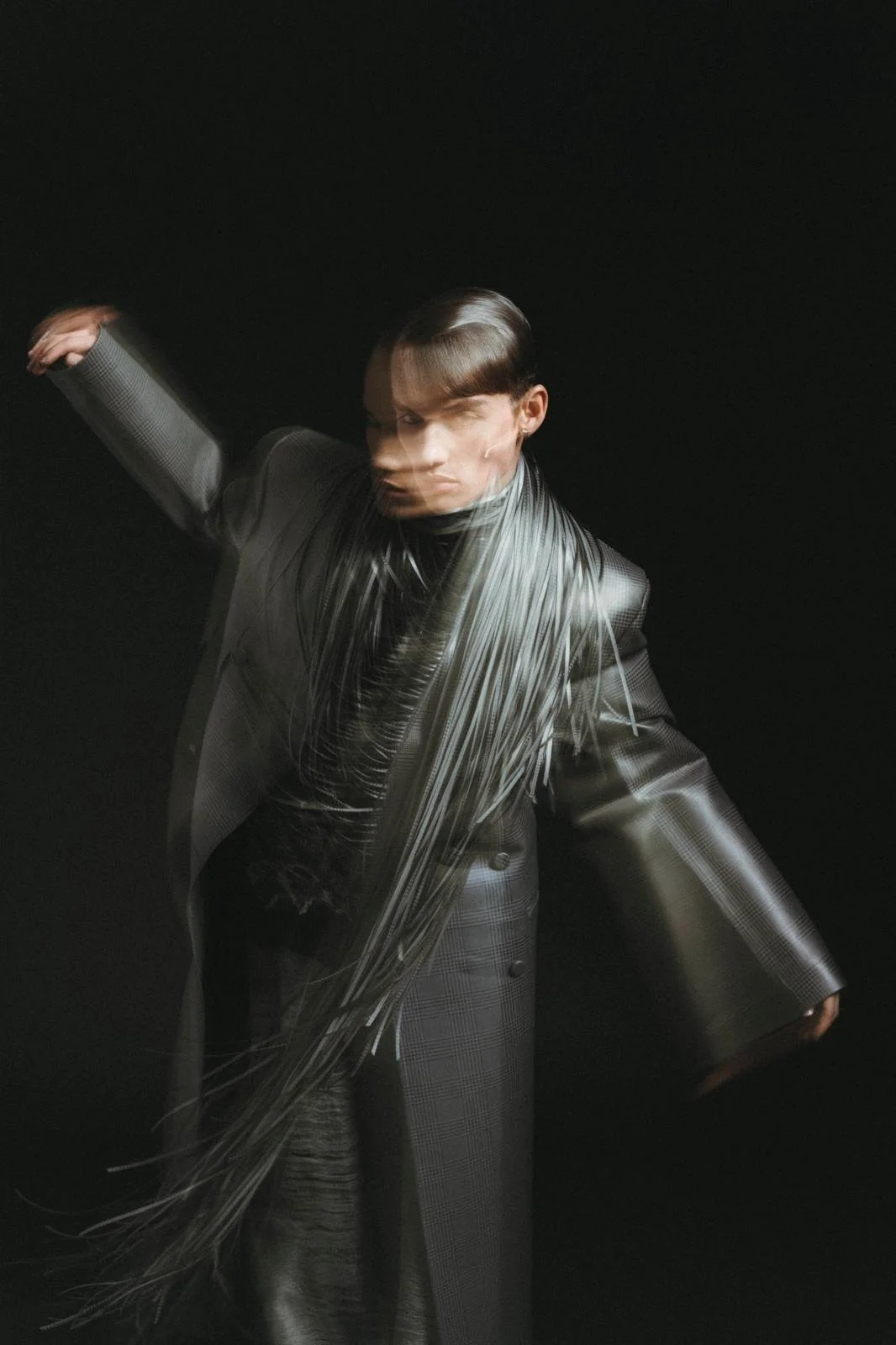 Artistic fashion photo of a person wearing a fringed scarf and a plaid coat, in motion blur, against a dark background.