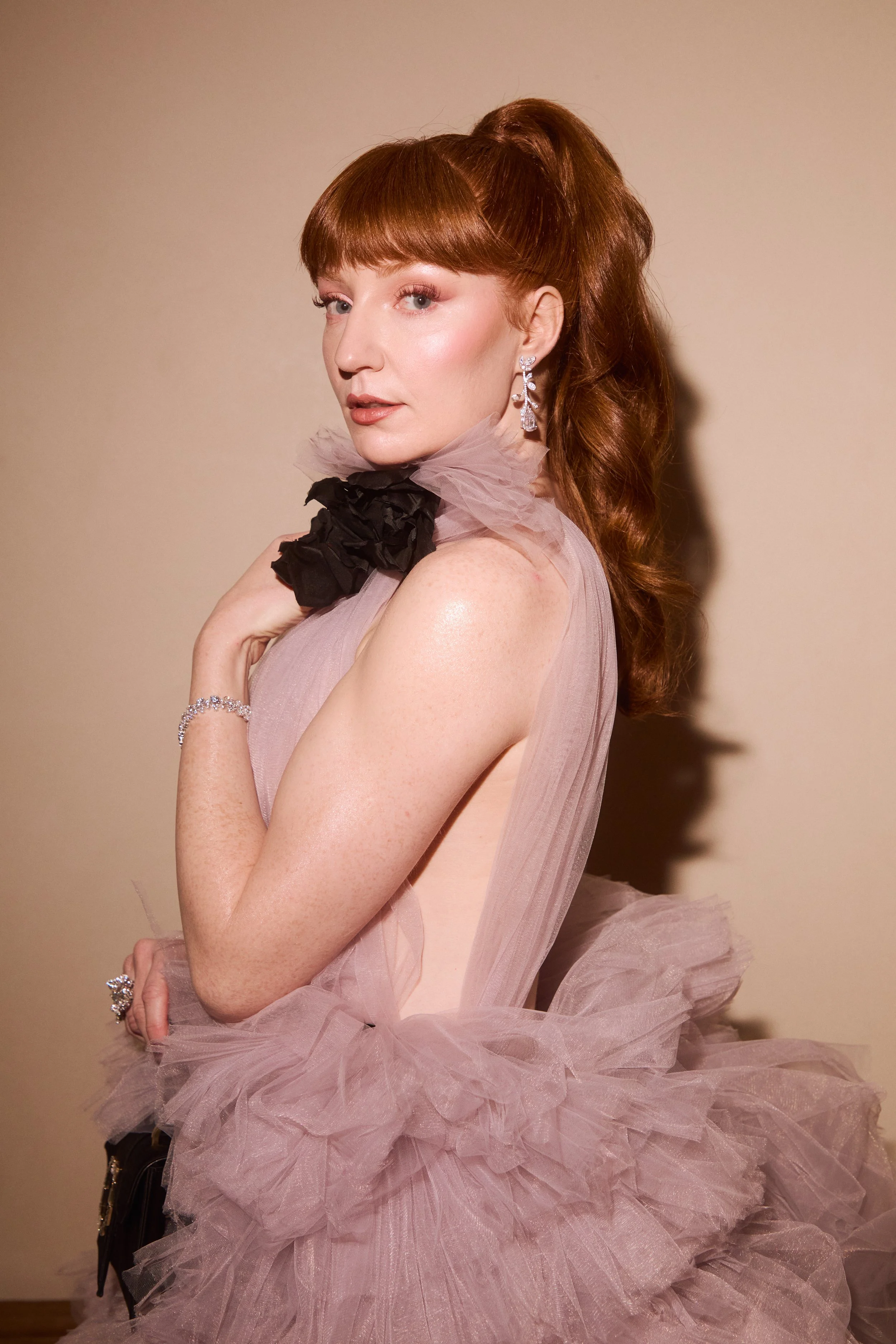Woman with red hair and a high ponytail wearing a ruffled mauve dress with black accents, posing against a neutral background.